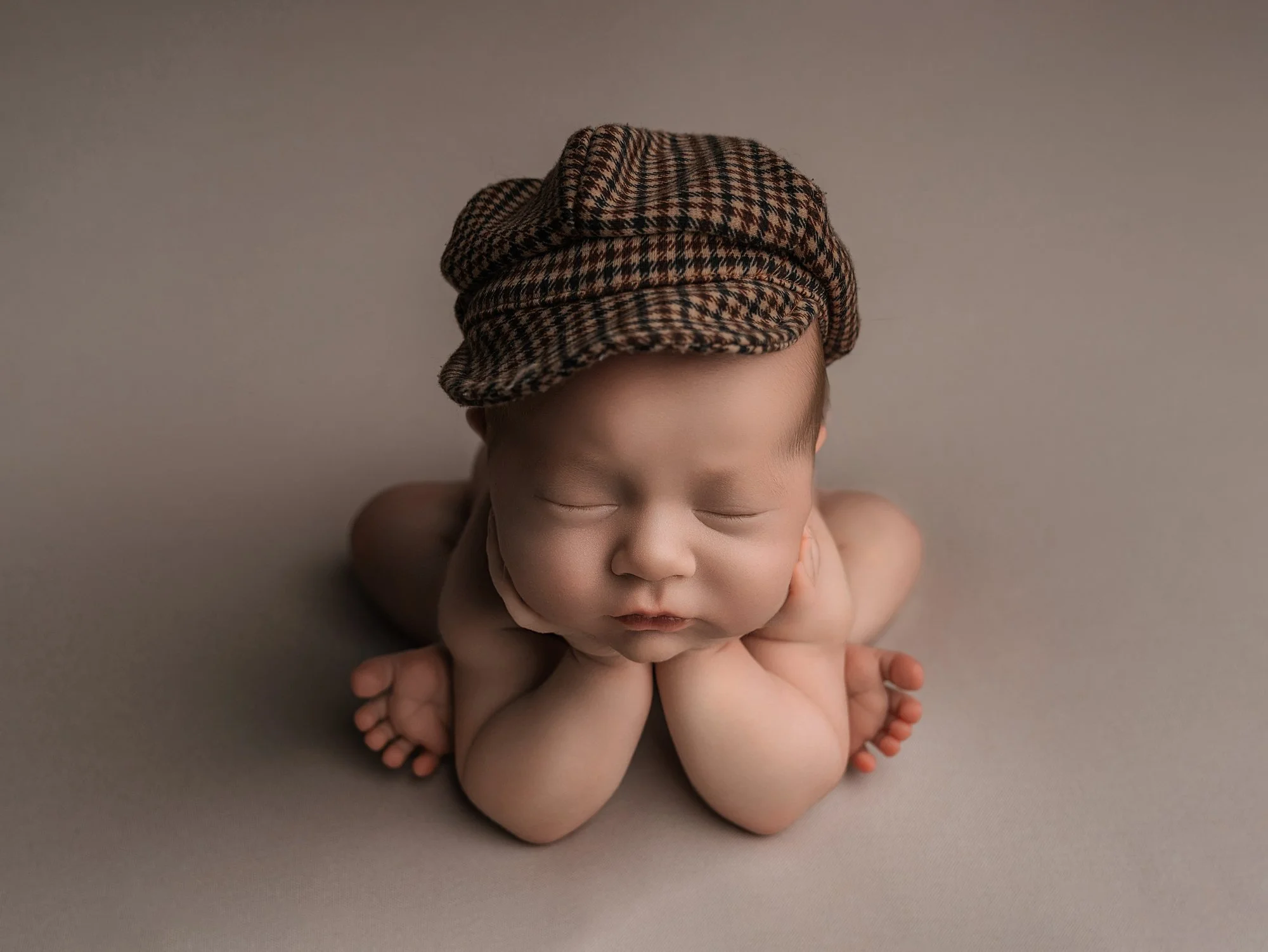 A sleeping baby wearing a brown plaid cap, lying on a soft neutral background with chin resting on hands.