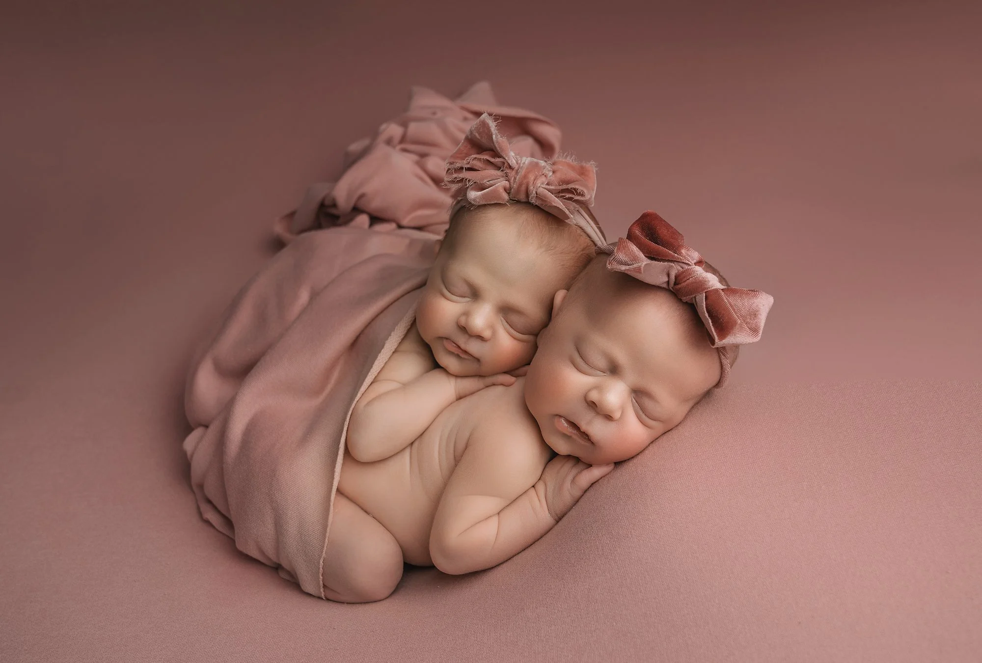 Two sleeping newborn babies, wrapped in a soft pink blanket and wearing matching pink headbands with bows, laying close together on a pink surface.