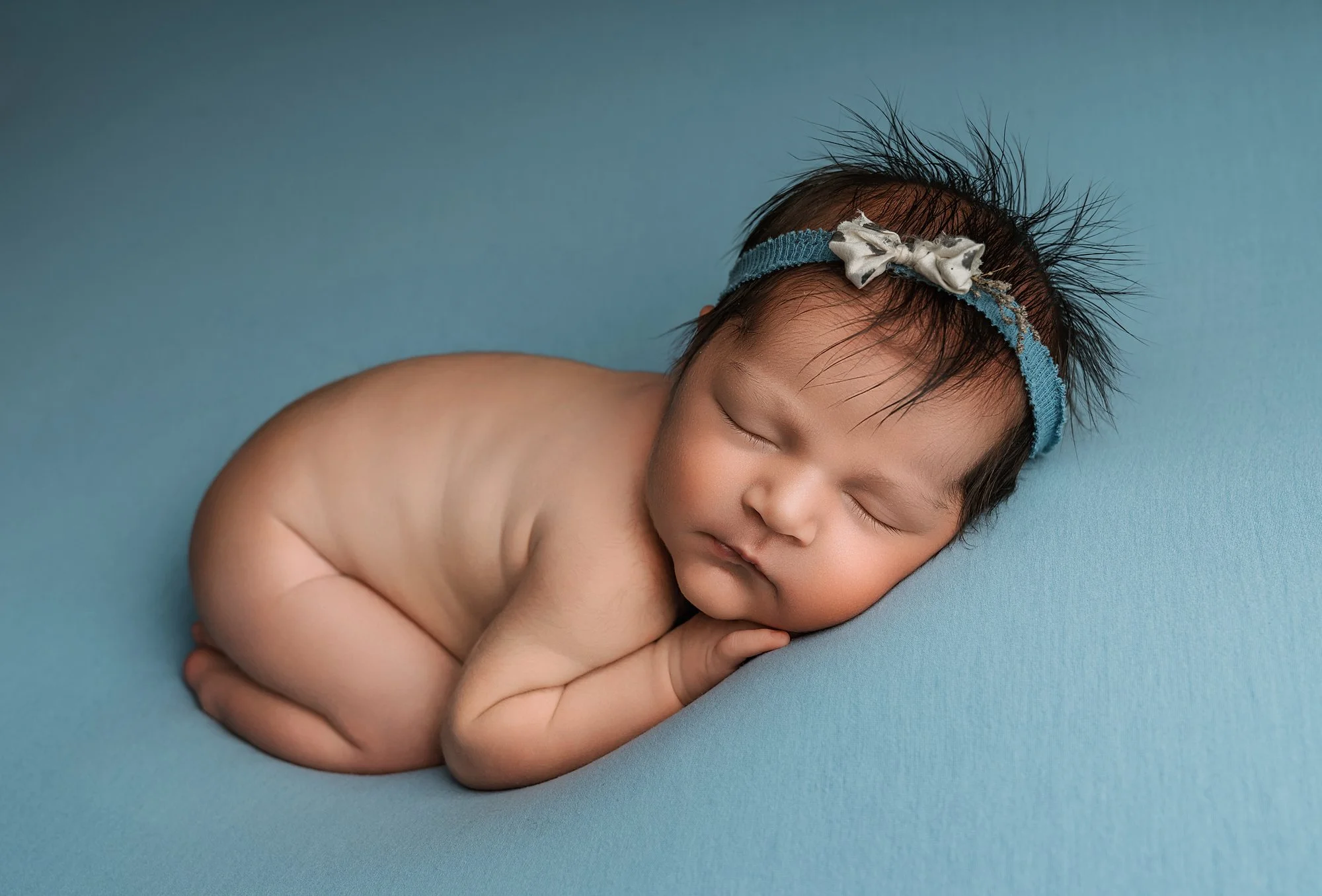 A sleeping baby with a blue headband and a small bow, lying on a blue surface, curled up on their side with hands under their cheek.