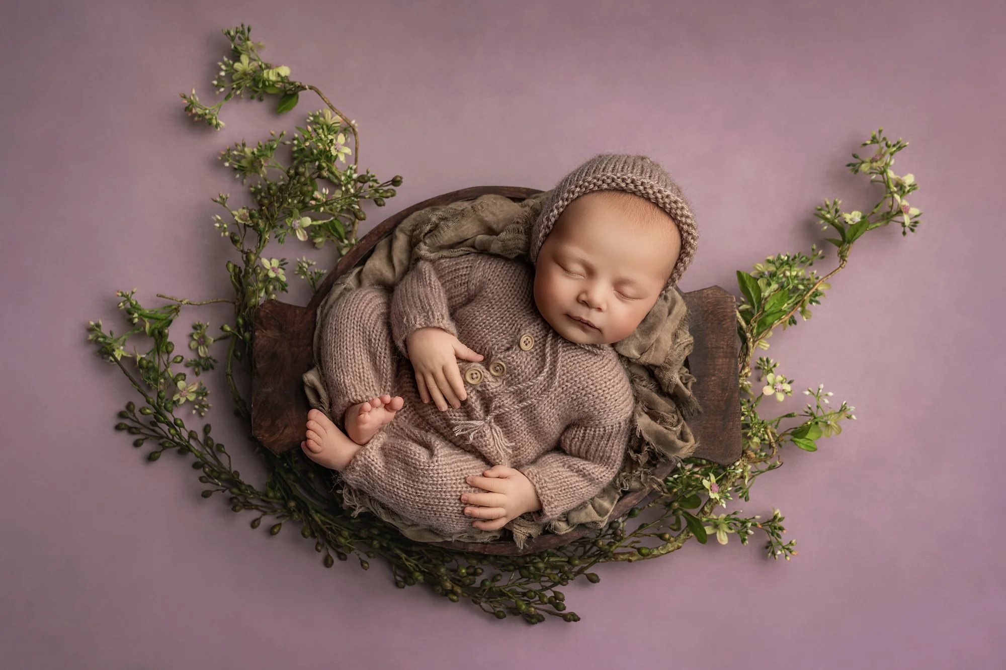 A sleeping baby in a knitted outfit and cap, lying curled up on a soft blanket, surrounded by decorative branches with small white flowers on a purple background.