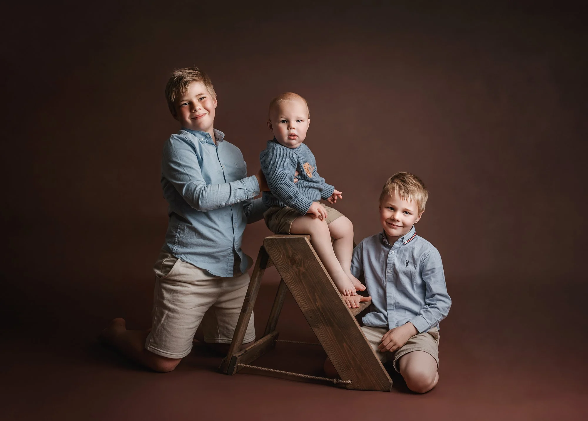 Three young boys with light hair, dressed in button-up shirts and shorts, posing with a young toddler boy wearing a blue sweater, sitting on a wooden ladder against a brown background. photographed at kath evans photography studio talgarth brecon mid