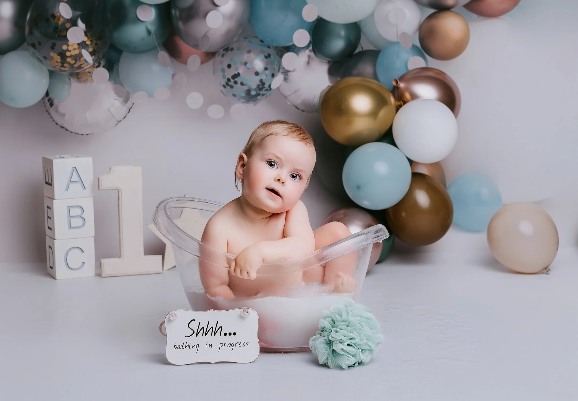 A baby sitting in a clear plastic tub filled with water, surrounded by balloons in shades of blue, gold, and white, photographed at kath evans photography studio talgarth brecon mid wales