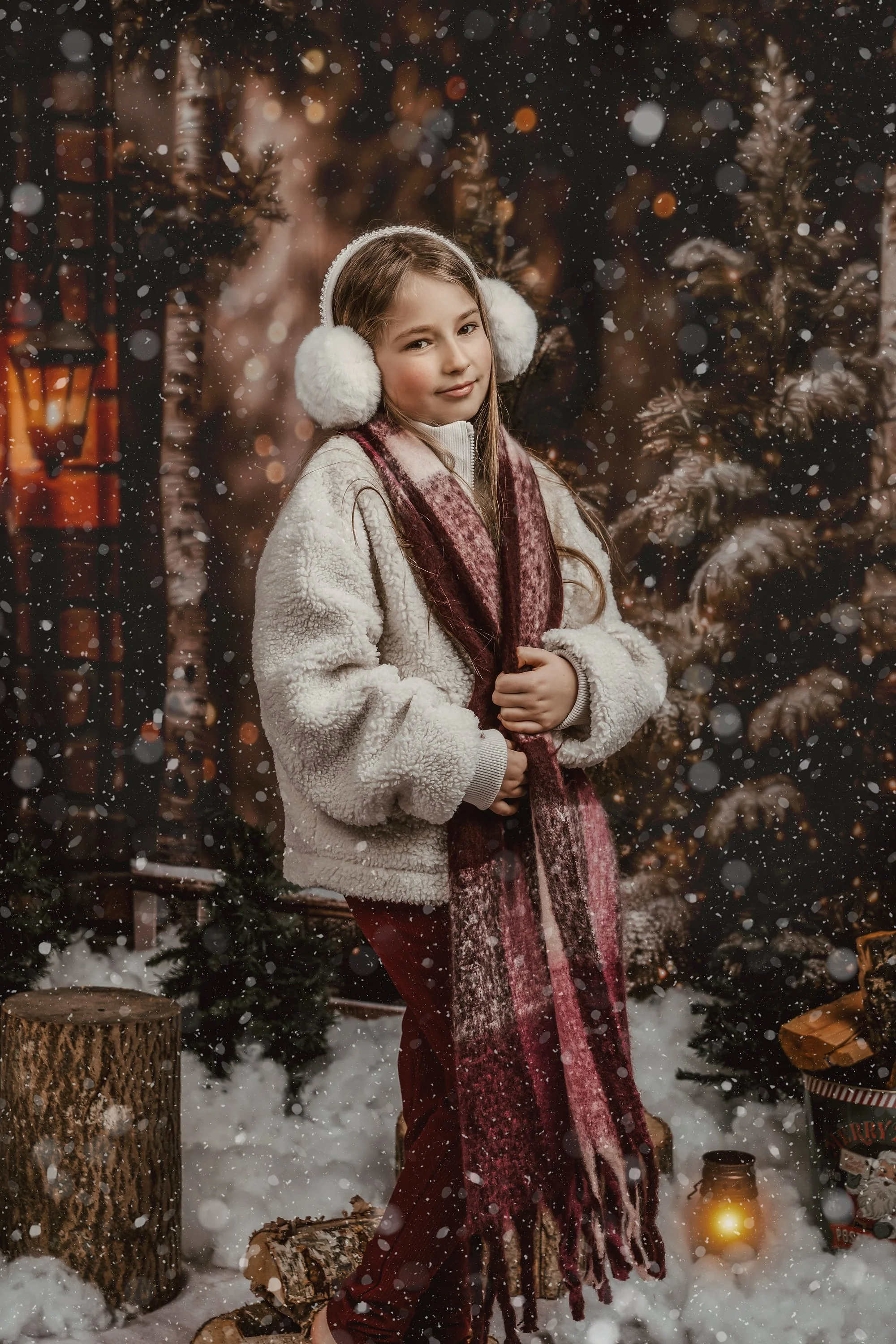 A young girl sitting on a brown chair in a Christmas decorated room, smiling at the camera. She wears a red dress with white shirt underneath. A stuffed teddy bear is on the floor next to her. The background features a decorated Christmas tree, toys, and holiday-themed decorations.