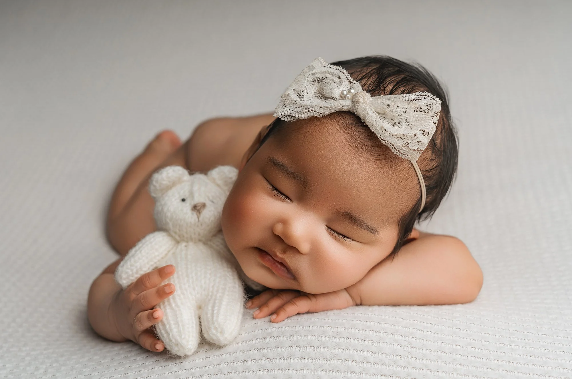 Young child with a lace bow headband sleeping on a white textured surface, hugging a knitted white teddy bear
