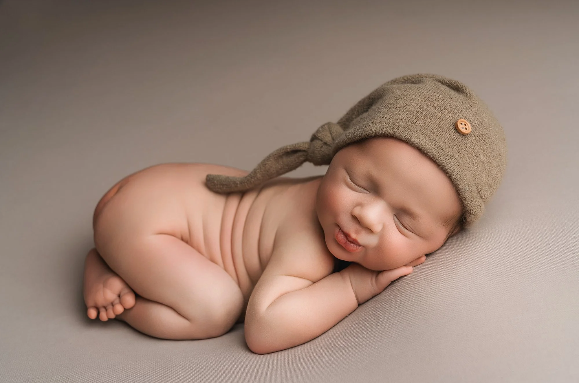 A sleeping newborn baby lying on its side on a soft surface, wearing a tan knit hat with a button, photographed at kath evans photography studio talgarth brecon mid wales.