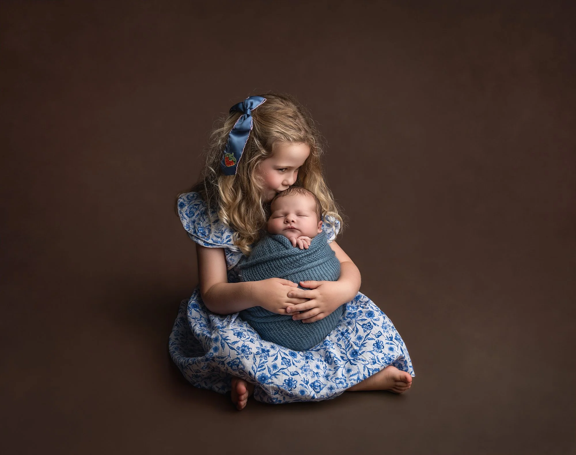 A young girl with long, curly blonde hair, wearing a blue and white floral dress and a blue headband, sitting on a brown background, holding a newborn baby wrapped in a blue blanket, with her head gently bowed over the baby's head.