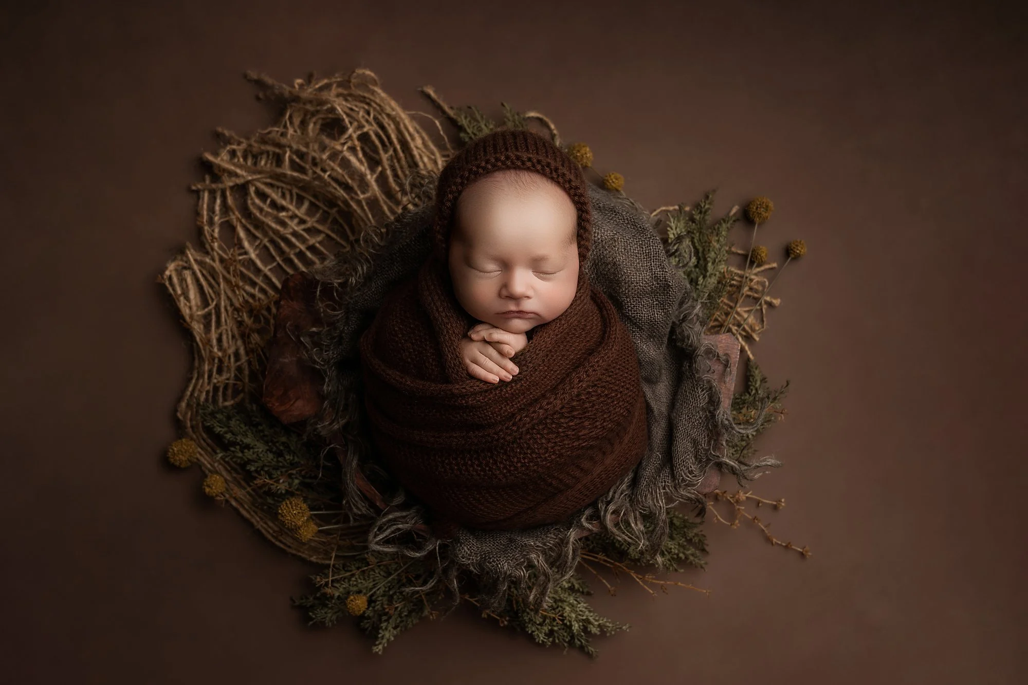 A peacefully sleeping newborn baby with a beige hat, resting with hands under chin inside a wooden bowl, surrounded by brown fabric on a wooden surface. photographed at kath evans photography studio talgarth brecon mid wales