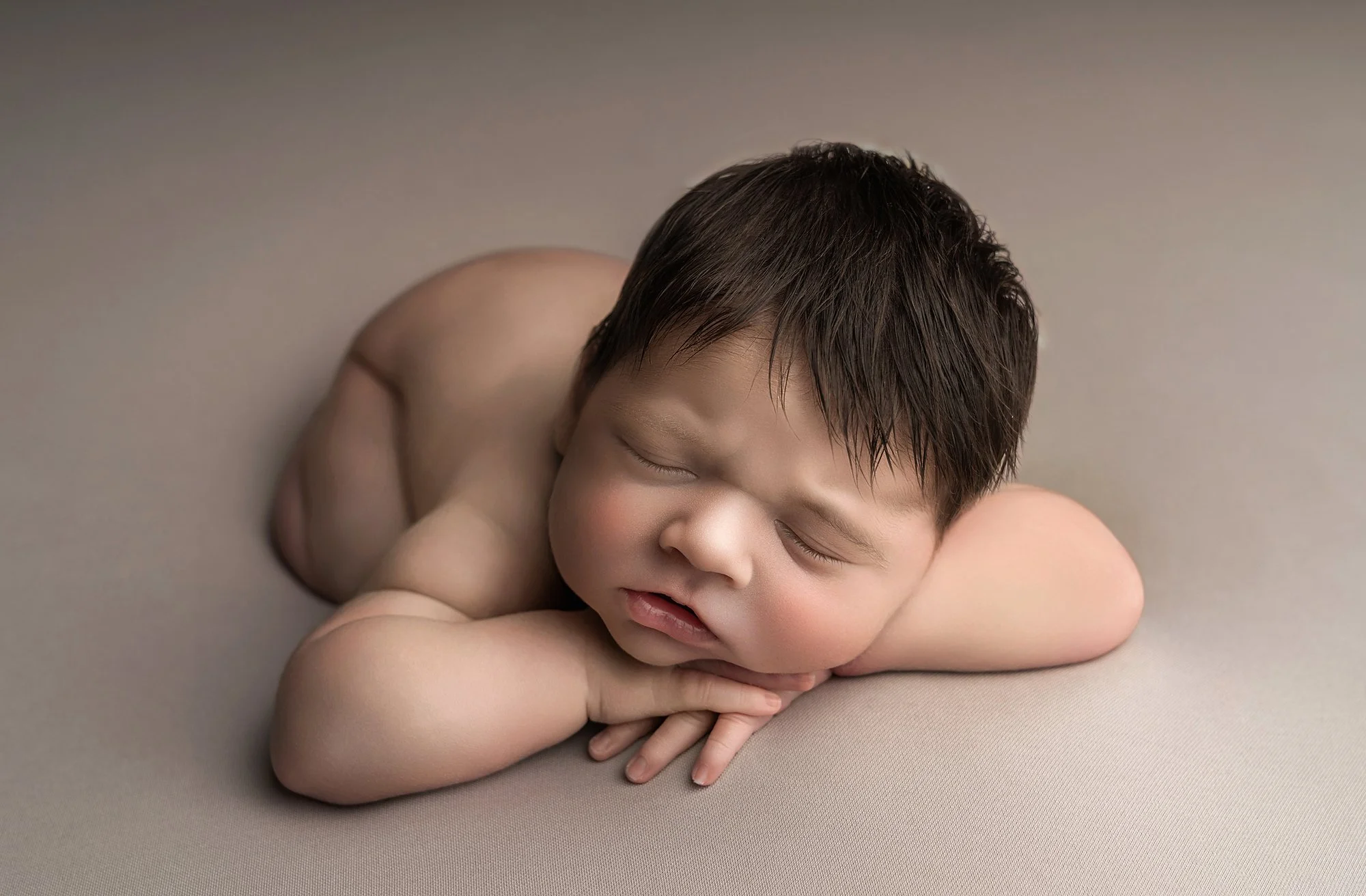 A young child with dark hair sleeping, laying on a beige surface with his arms crossed under his chin.