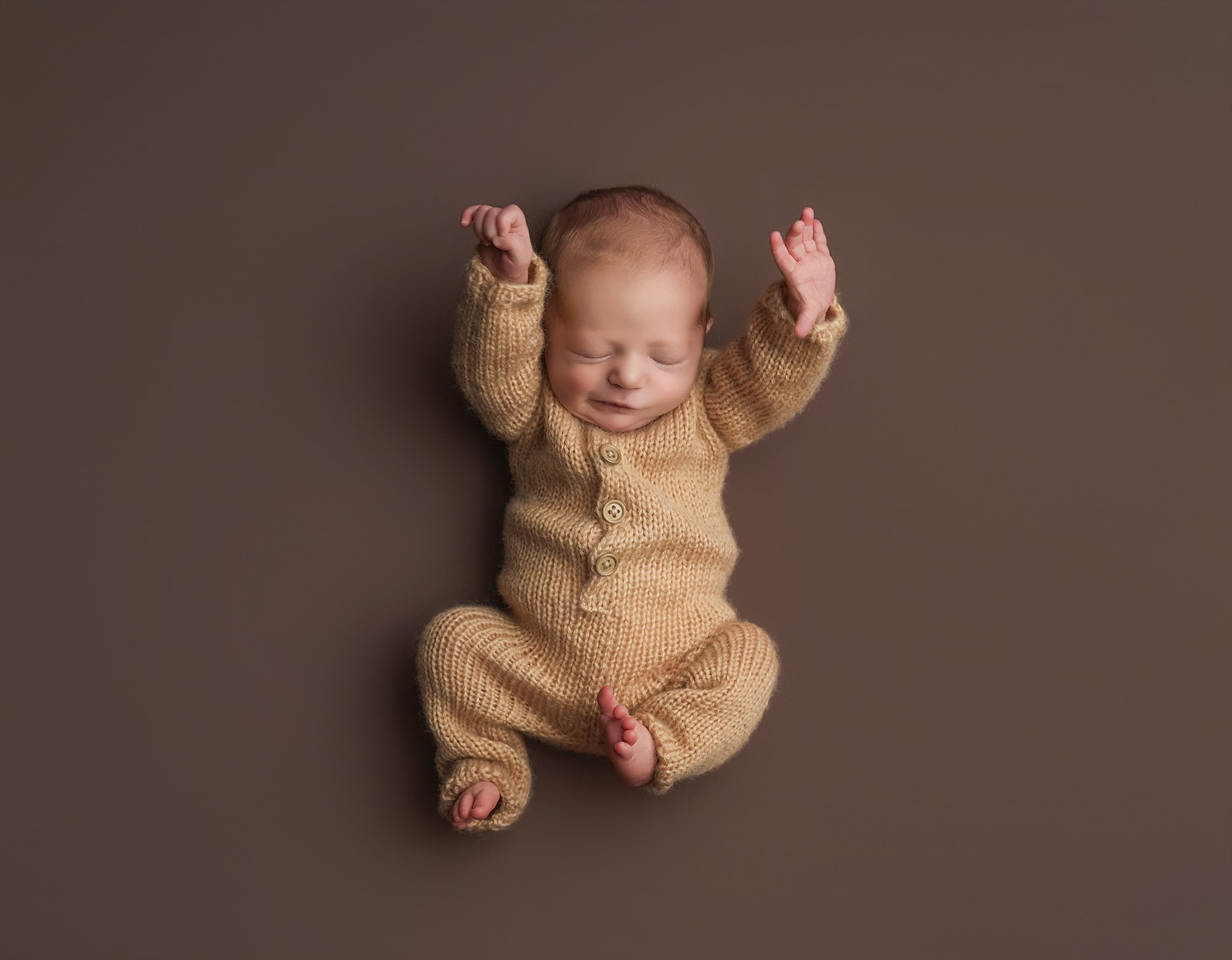 A sleeping newborn baby in a knitted tan onesie lying on a brown surface with arms raised above head and a peaceful expression.