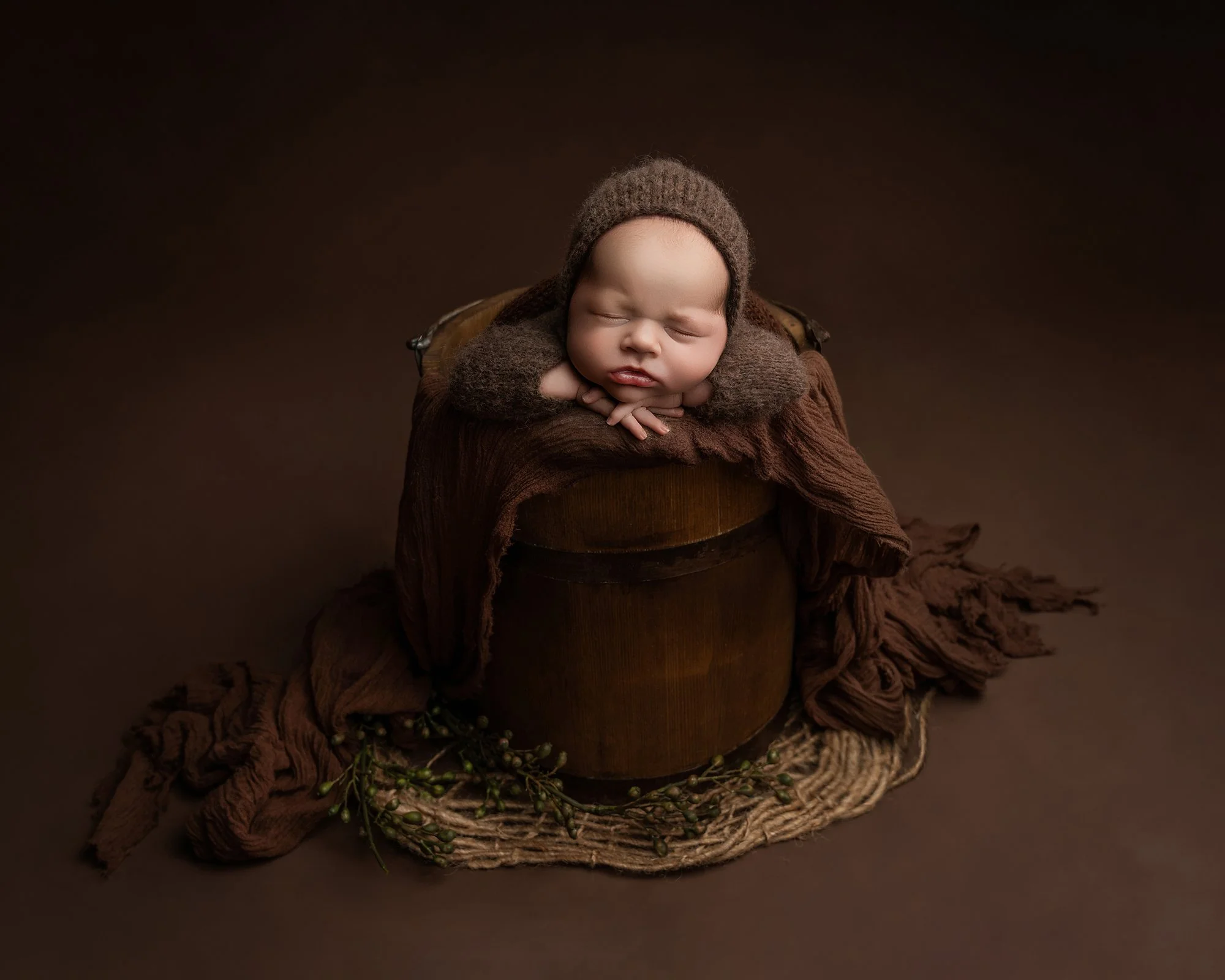 Cute baby sleeping with closed eyes, resting on a brown wooden box, adorned in a knitted brown hat and matching outfit, surrounded by dark brown and tan fabric, with small green sprigs at the base, set against a dark background.