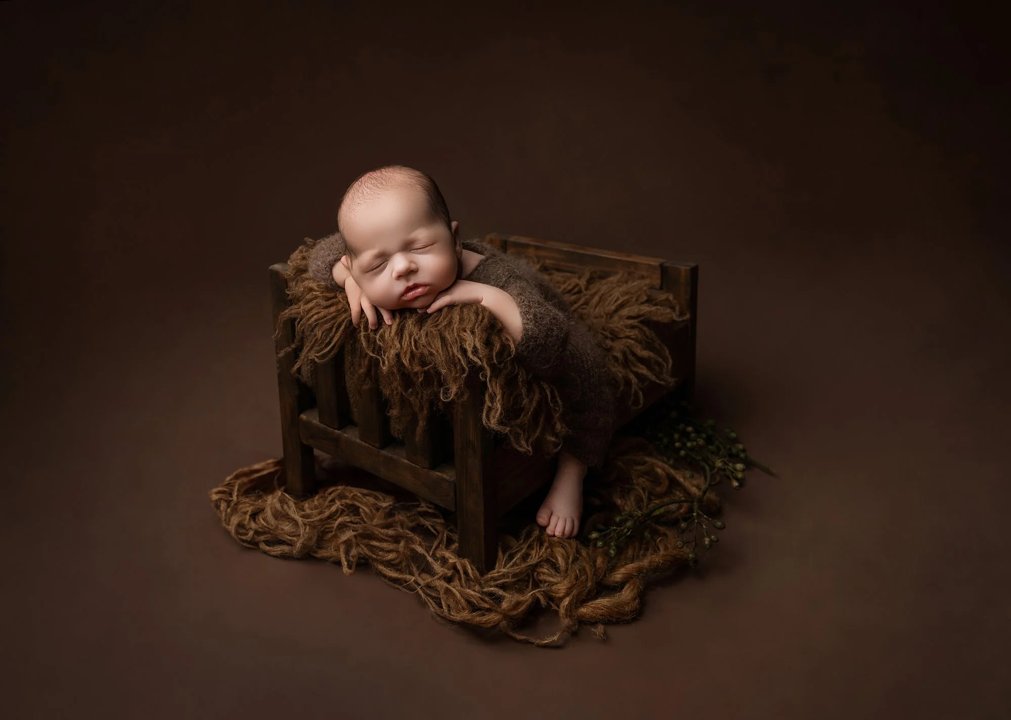 A newborn baby sleeping on a small wooden bed with brown cozy blankets and a brown background.
