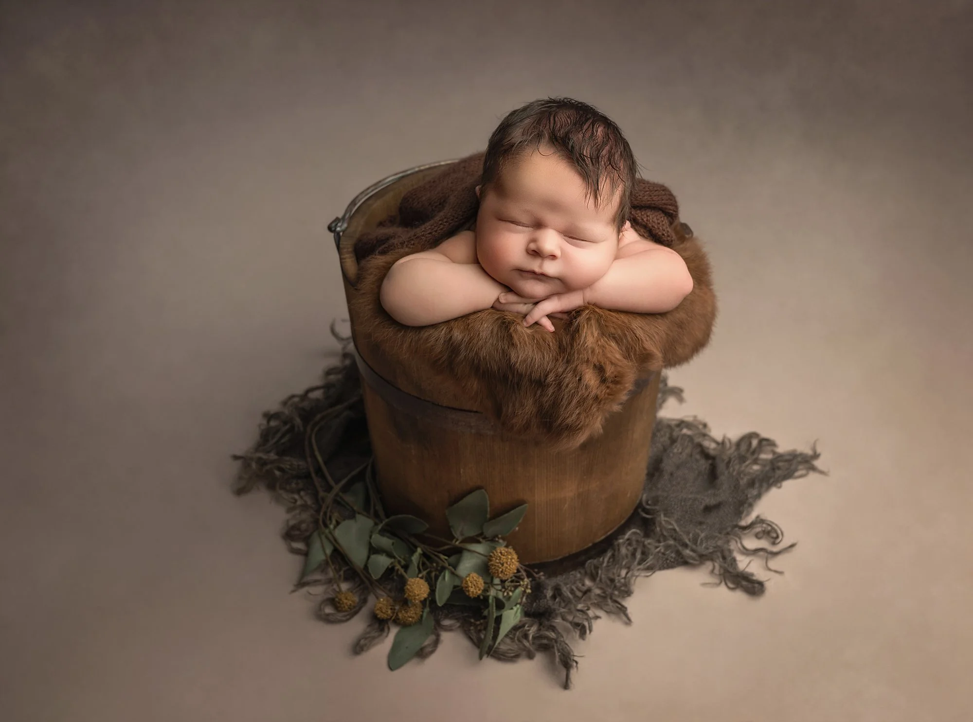 A newborn baby sleeping with arms folded under chin in a wooden basket lined with brown fur and a dark blanket, surrounded by green leaves and small yellow flowers on a wool rug.