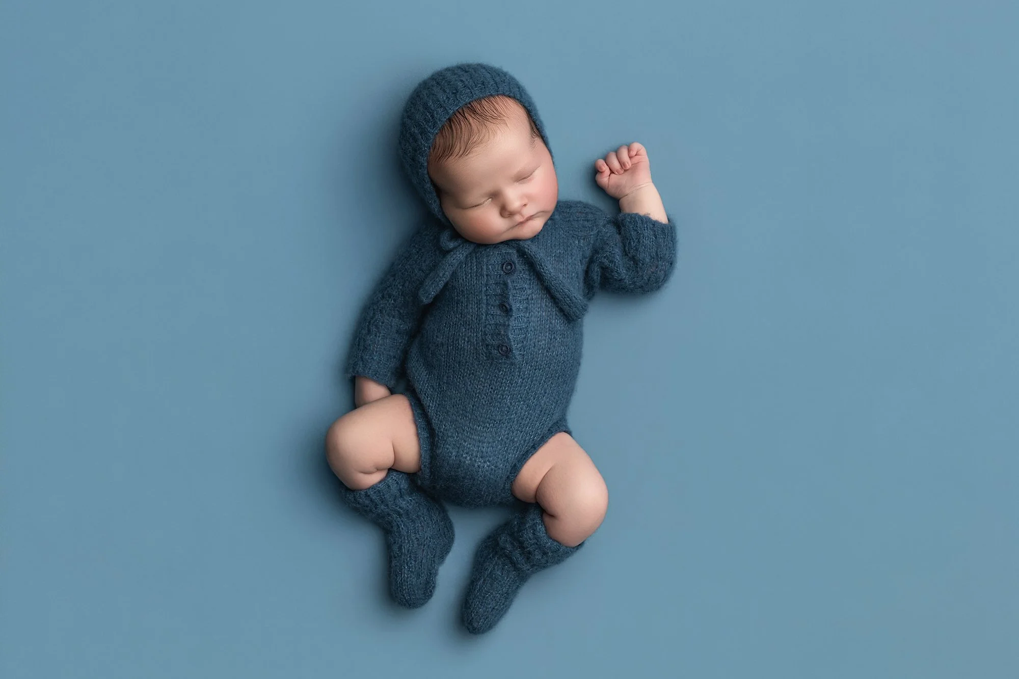 a newborn boy sleeping peacefully in a blue knitted romper on a blue blanket photographed at kath evans photography studio talgarth brecon mid wales