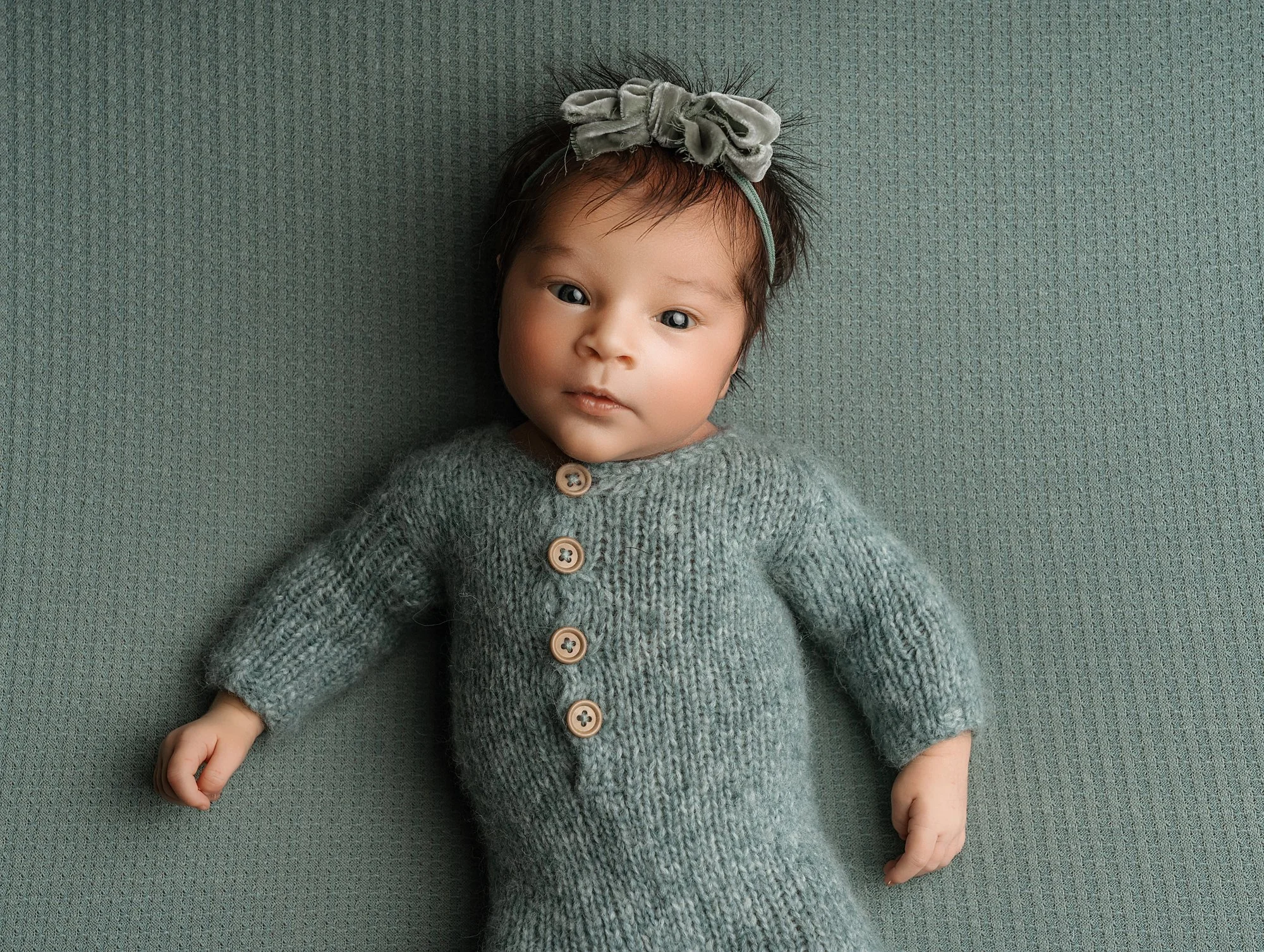 A baby girl with dark hair and blue eyes lying on a textured green surface, wearing a grey knitted outfit with wooden buttons and a grey headband with a bow. photographed at kath evans photography studio talgarth brecon mid wales