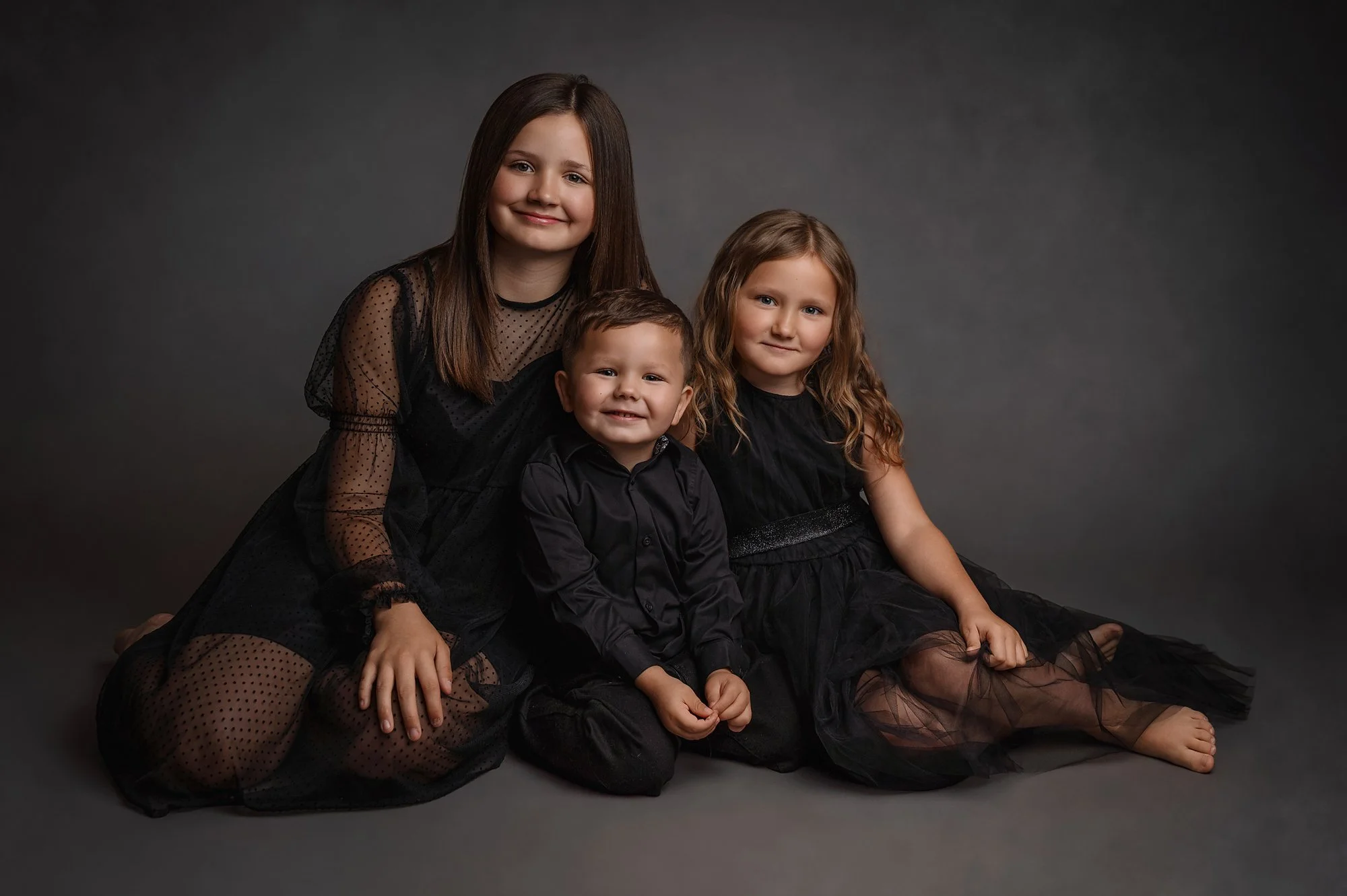 Three children, two girls and one boy, dressed in black, sitting against a dark gray background. photographed at kath evans photography studio talgarth brecon mid wales