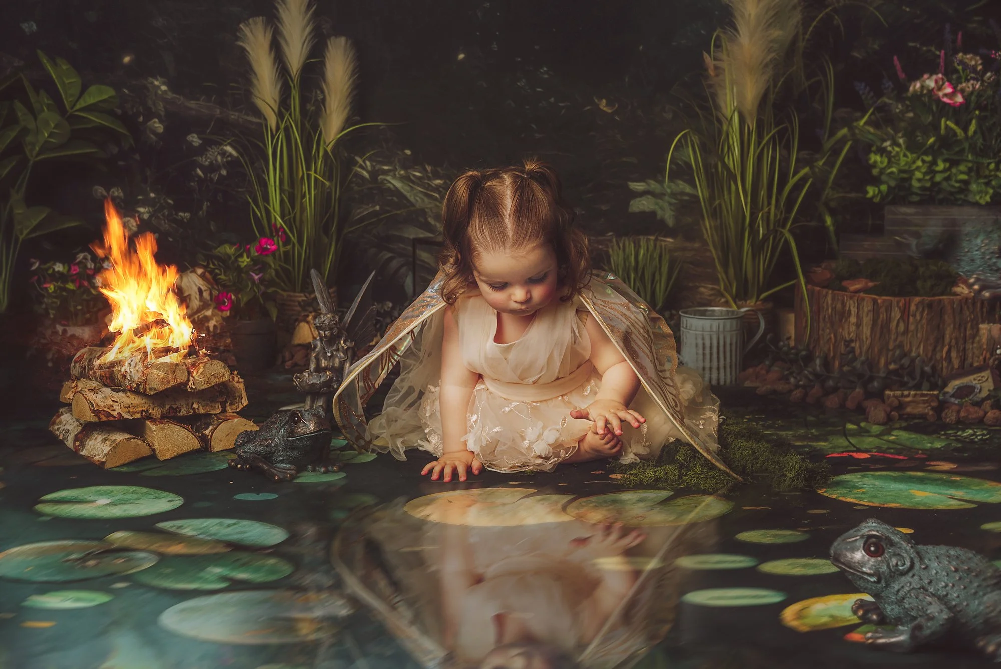 A young girl dressed as a fairy with wings, sitting by a pond with lily pads and frogs, surrounded by plants and a small fire on logs in a garden setting.