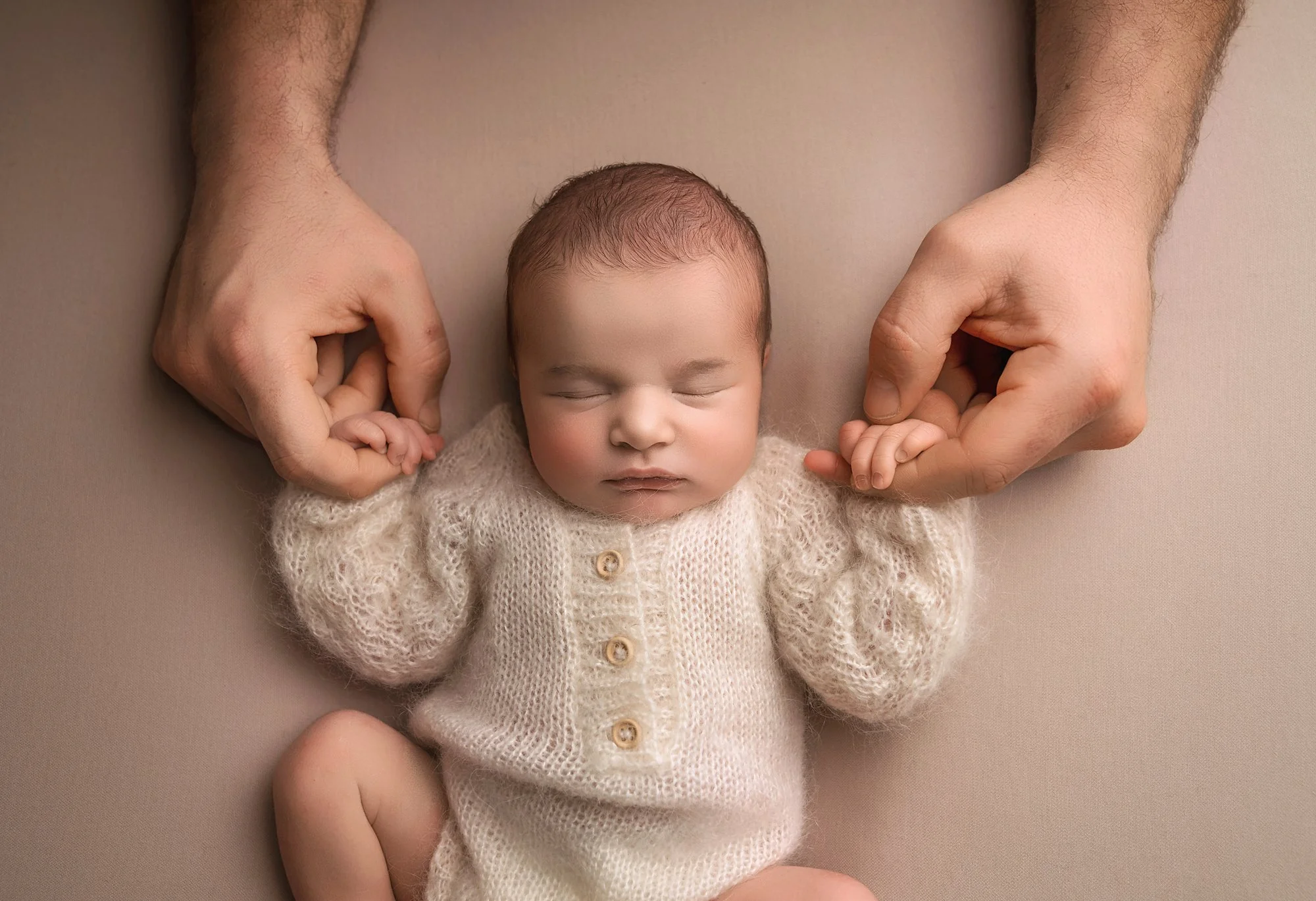 A newborn baby in a cream-colored knitted sweater sleeping with eyes closed, held gently by hands at the shoulders, lying on a neutral surface.
