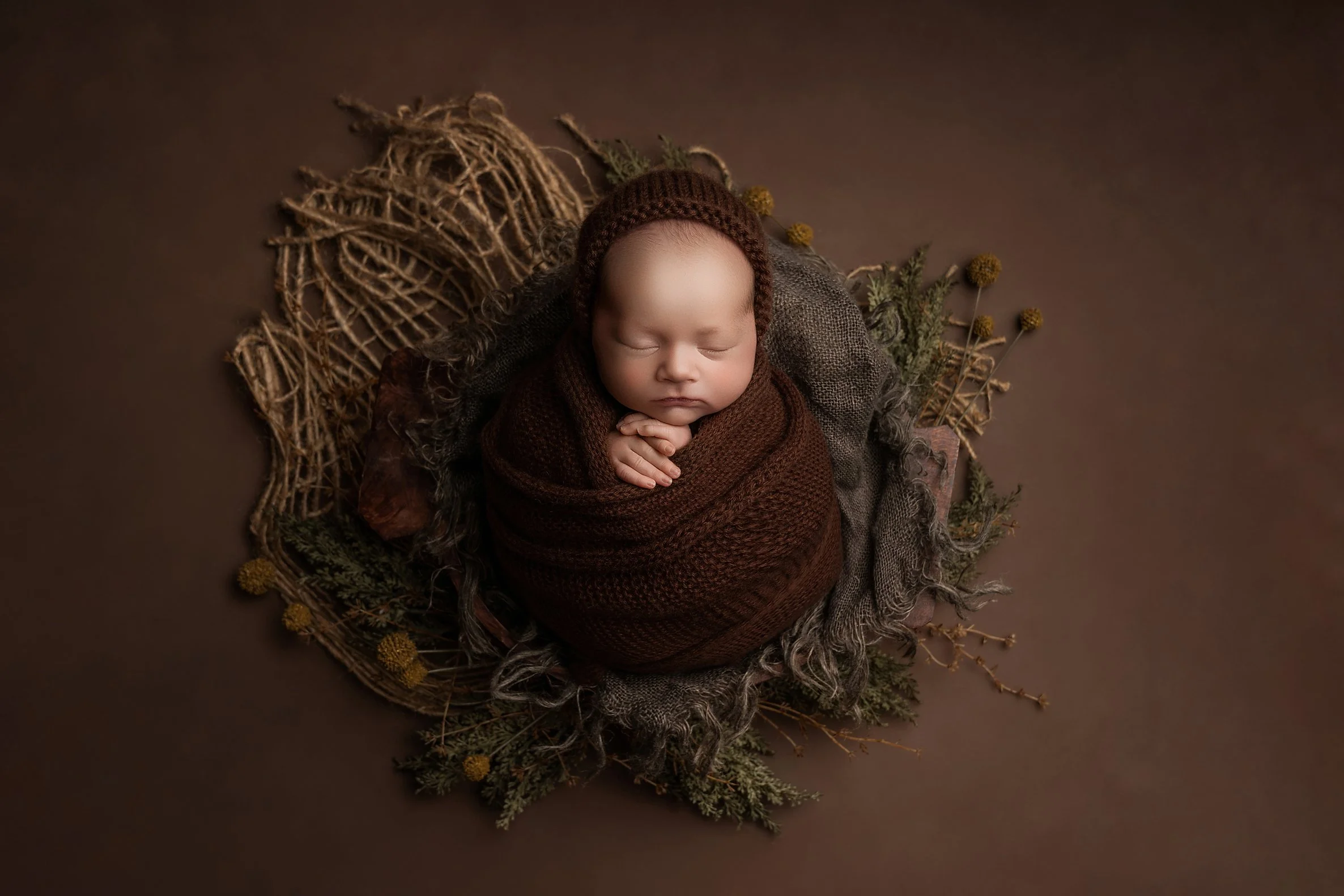 A newborn baby swaddled in a brown blanket, wearing a matching knit hat, sleeping peacefully on a brown background with rustic decorations around.