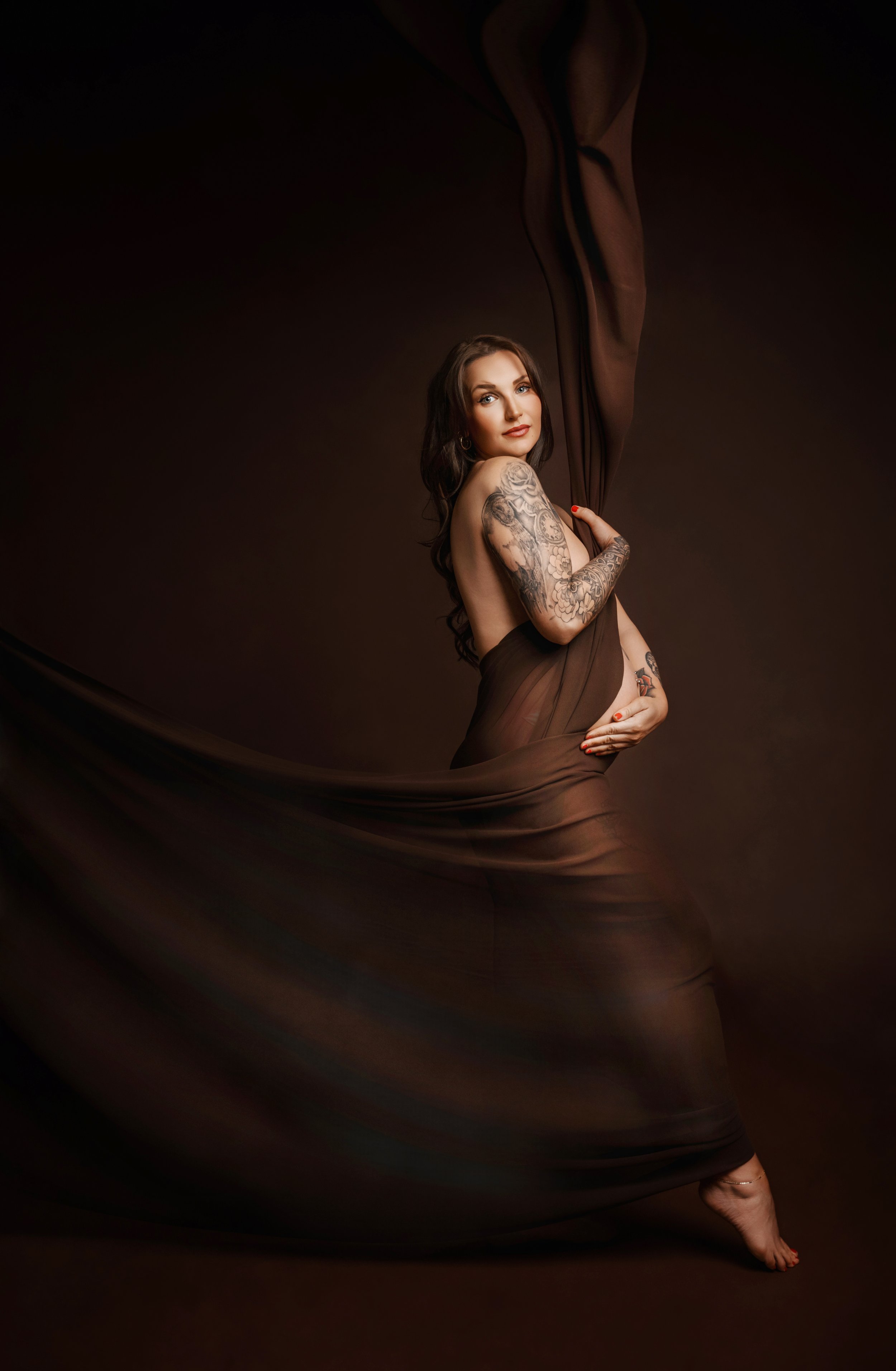 A pregnant woman with long dark hair and tattoos on her arms is posing with a flowing brown fabric against a dark background. photographed at kath evans photography studio talgarth brecon mid wales