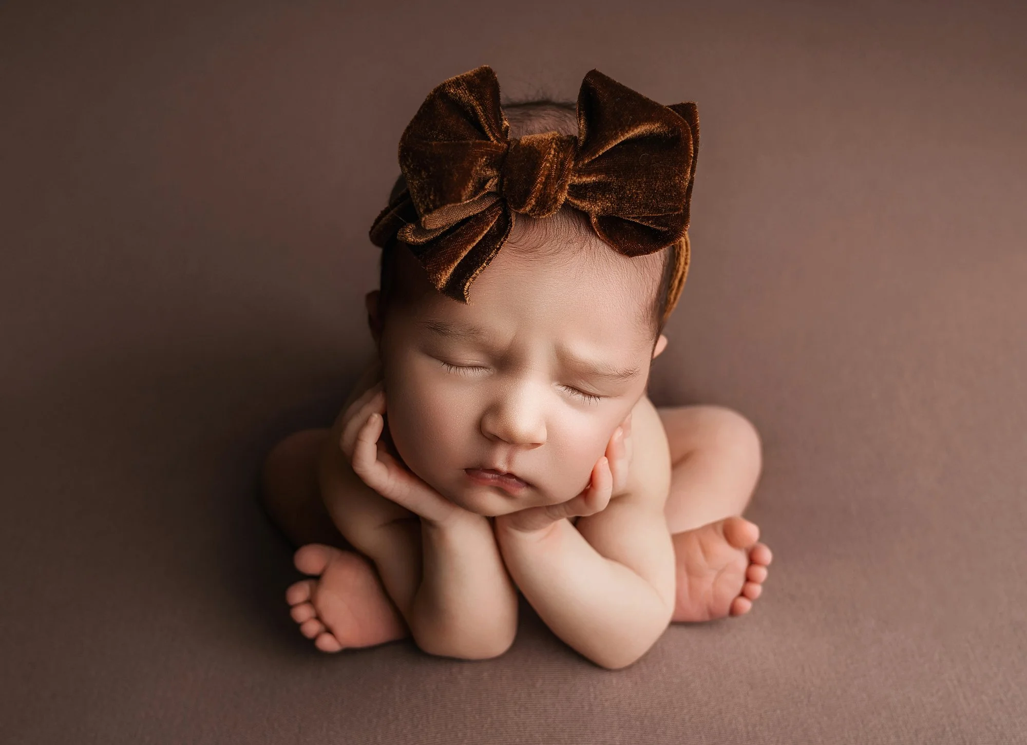 A sleeping baby girl with a brown velvet bow headband, resting her chin on her hands, sitting on a brown surface.