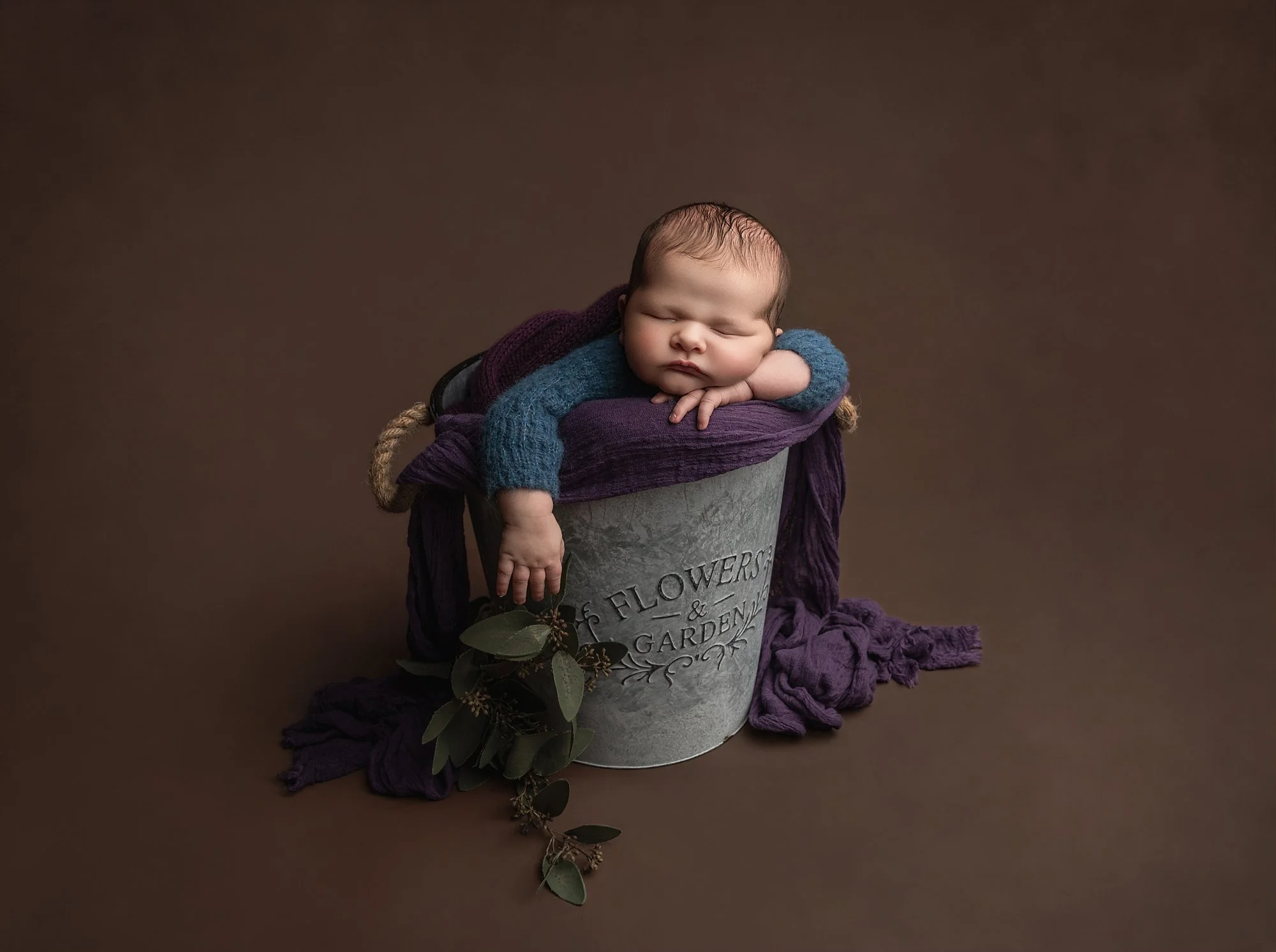 A sleeping baby with a blue sweater laying headrest on the arm, inside a silver flower and garden bucket, surrounded by purple and dark fabric, against a brown background.