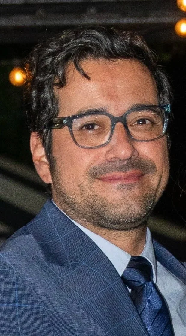 A man with dark hair and glasses, wearing a blue plaid suit and tie, smiling at a formal event with blurred lights in the background.