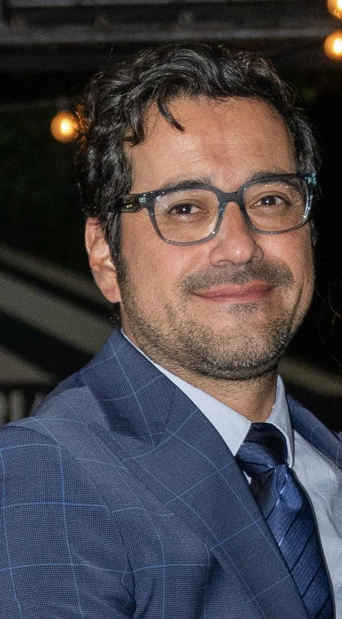 A man with dark, curly hair, glasses, and a beard, wearing a blue checkered suit and tie, smiling in a close-up portrait.
