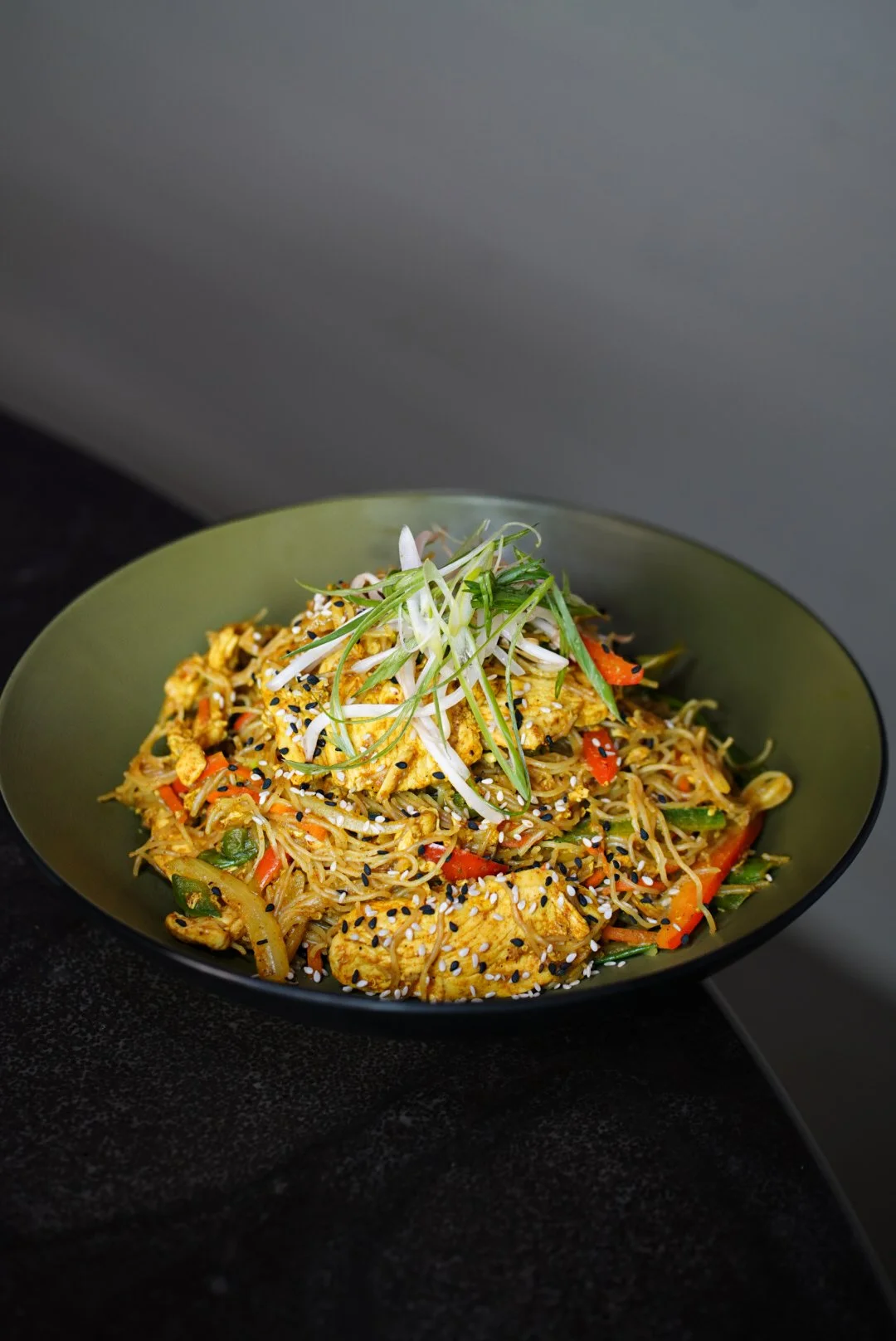 A bowl of Asian-style stir-fried noodles with vegetables and sliced chicken, garnished with chopped green onions and sesame seeds.