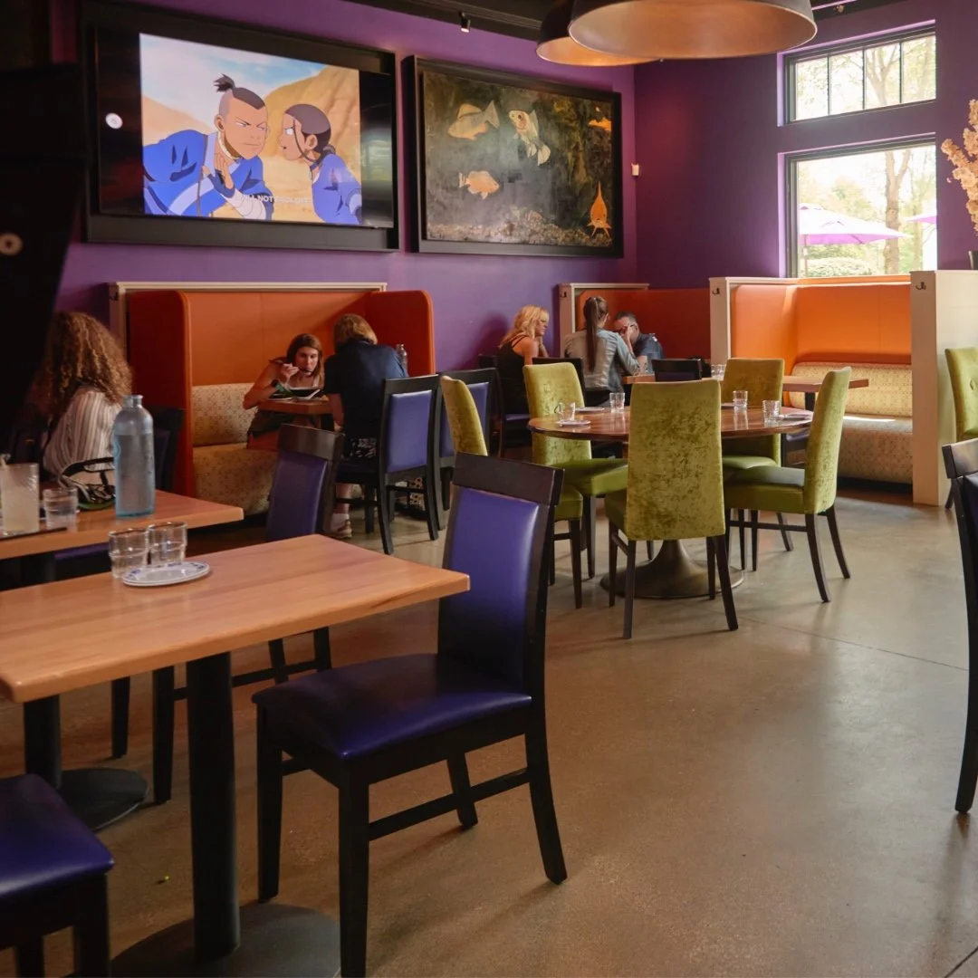 Modern Asian-inspired dining room with purple walls, colorful booths, and a large screen playing an animated show.