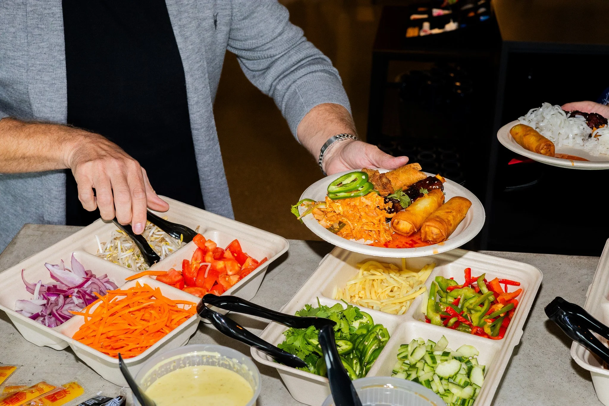 A person serving a plate of food with spring rolls, shredded carrots, green chili peppers, and other vegetables at a buffet-style setup with various vegetables in bowls.