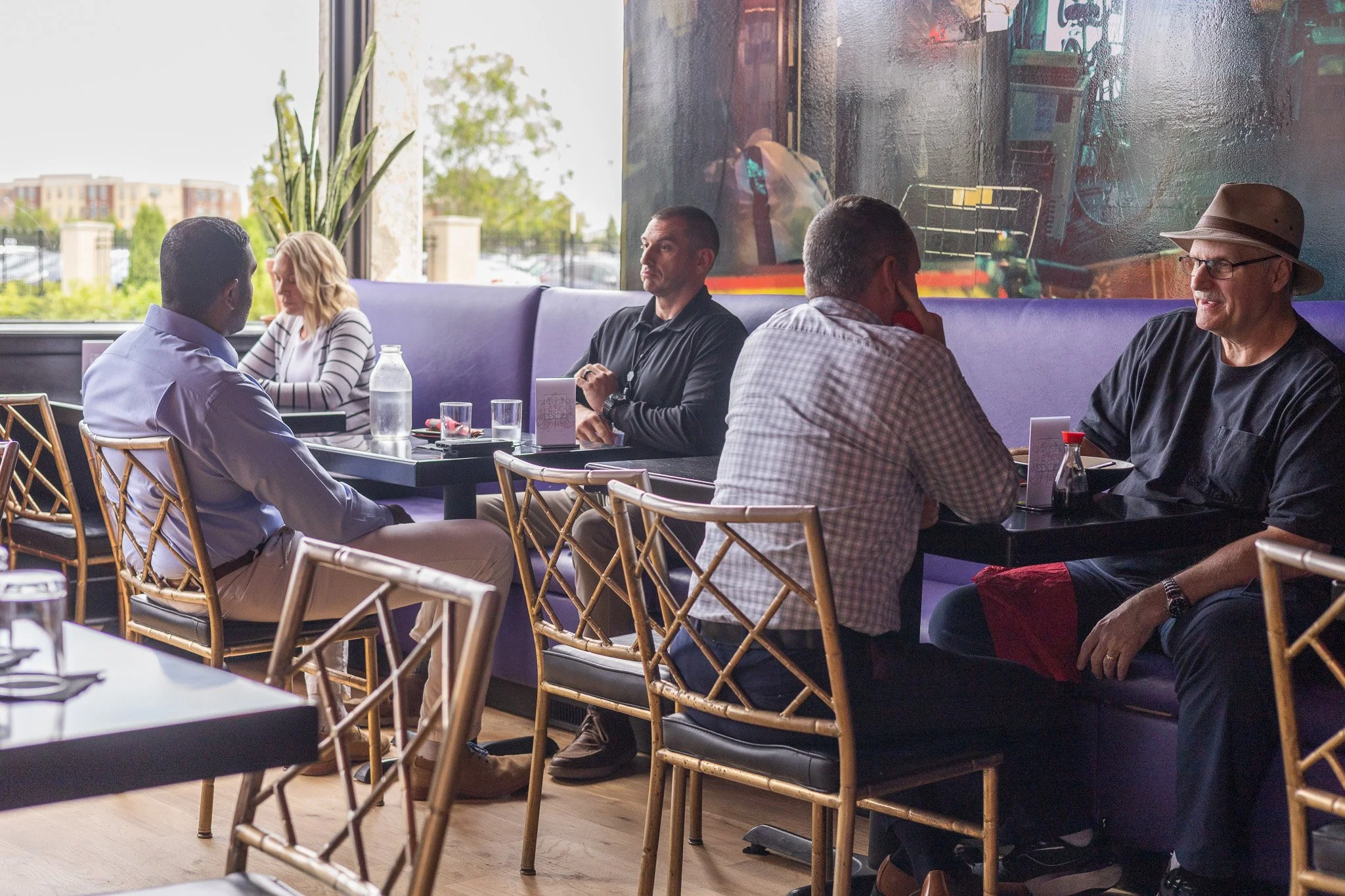 People sitting at tables in a restaurant with large windows, a purple booth along the window, and a colorful abstract painting on the wall.