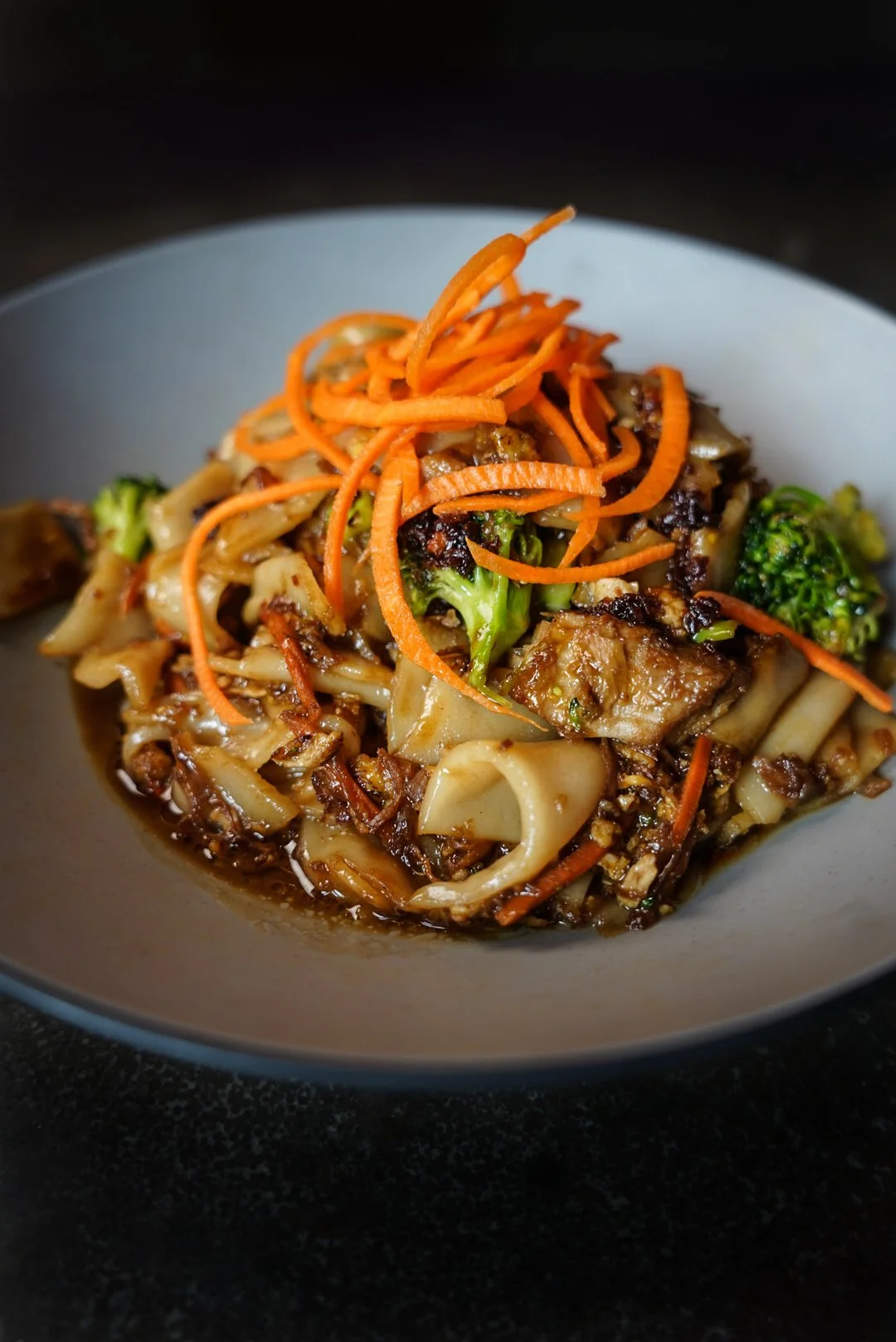 A bowl of stir-fried noodles with vegetables including broccoli, carrots, and onions, garnished with shredded carrots.