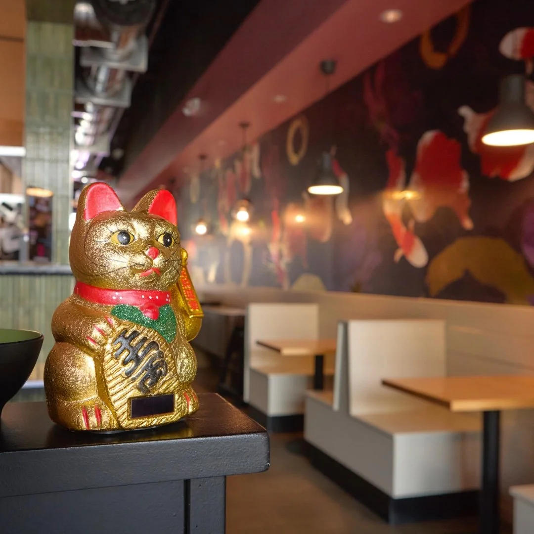 Golden Maneki-neko lucky cat statue displayed at the entrance of a restaurant with booth seating and floral mural in the background.