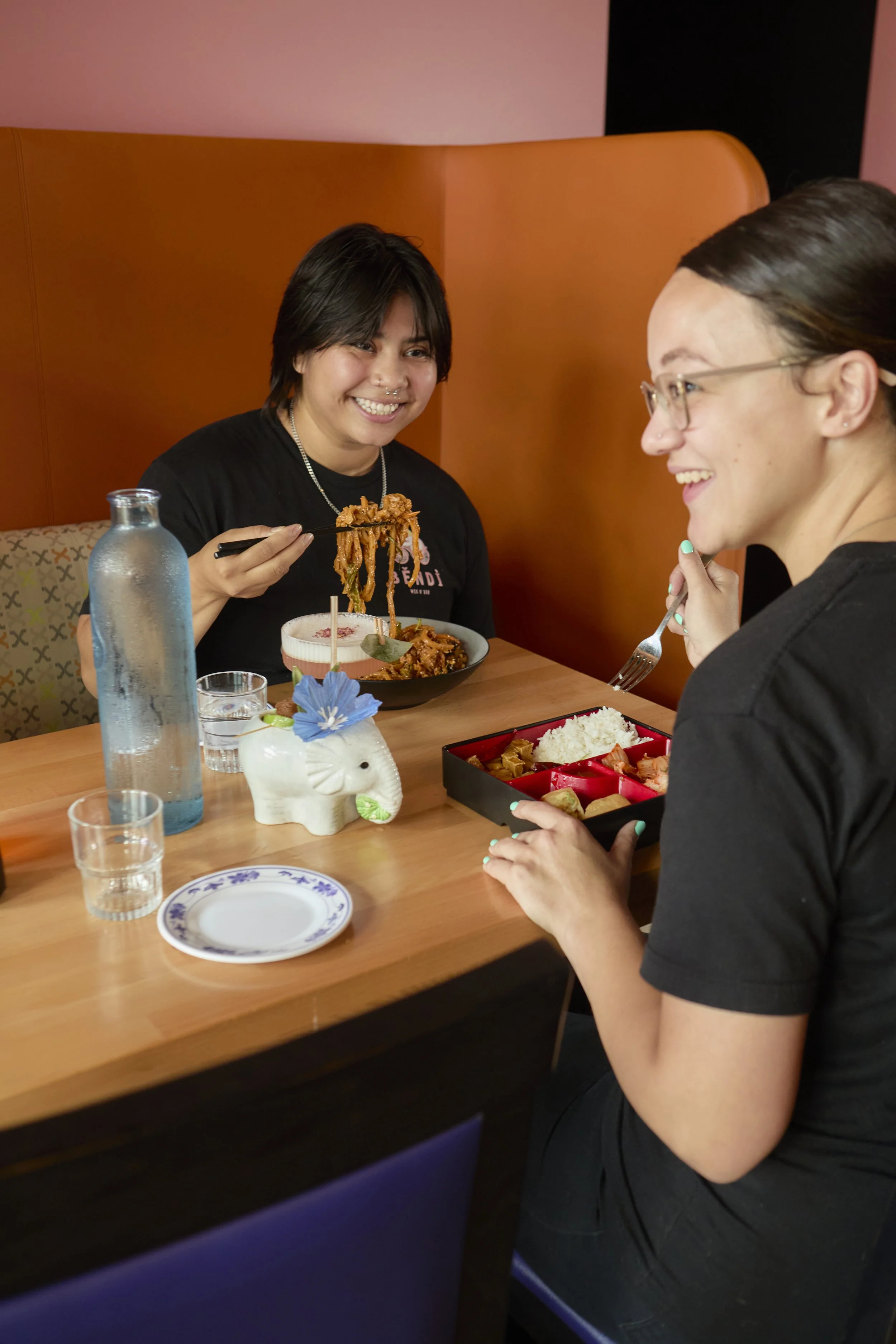 Two women sitting at a restaurant table, smiling while sharing a meal of noodles and rice. One woman has dark hair and glasses, the other has short dark hair and a nose piercing. The table has a water bottle, a small flower planter, and dishes of food.