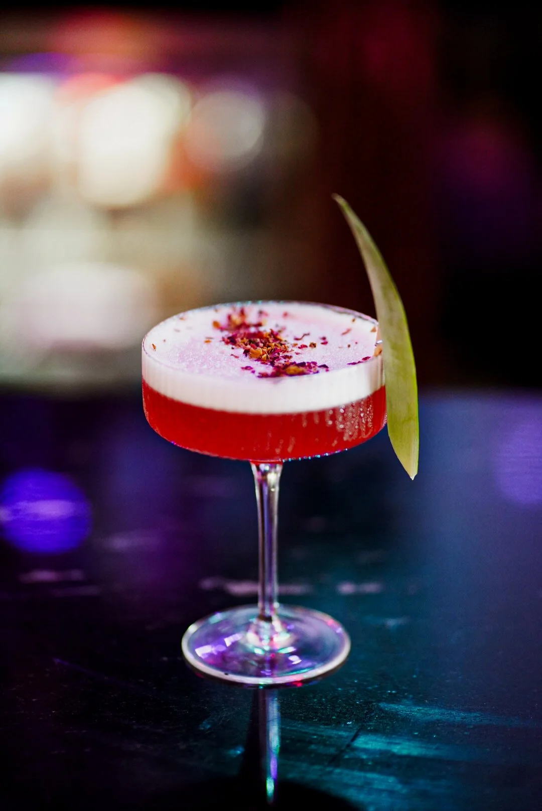 A fancy cocktail in a coupe glass with a red bottom layer and a white frothy top, garnished with a green leaf and pink flower petal decorations on top, set against a blurred colorful background.
