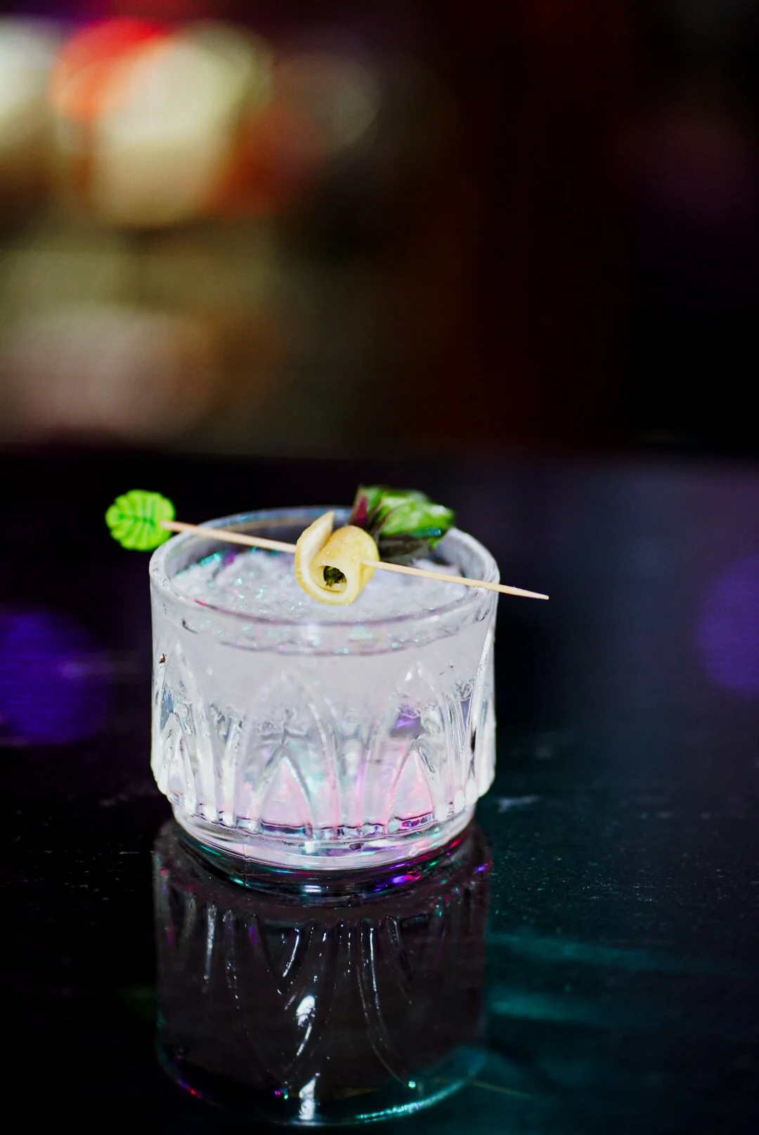 A glass of clear cocktail with ice, garnished with a lemon twist, a small green decorative pick, and a purple flower, on a dark reflective surface with a blurred colorful background.
