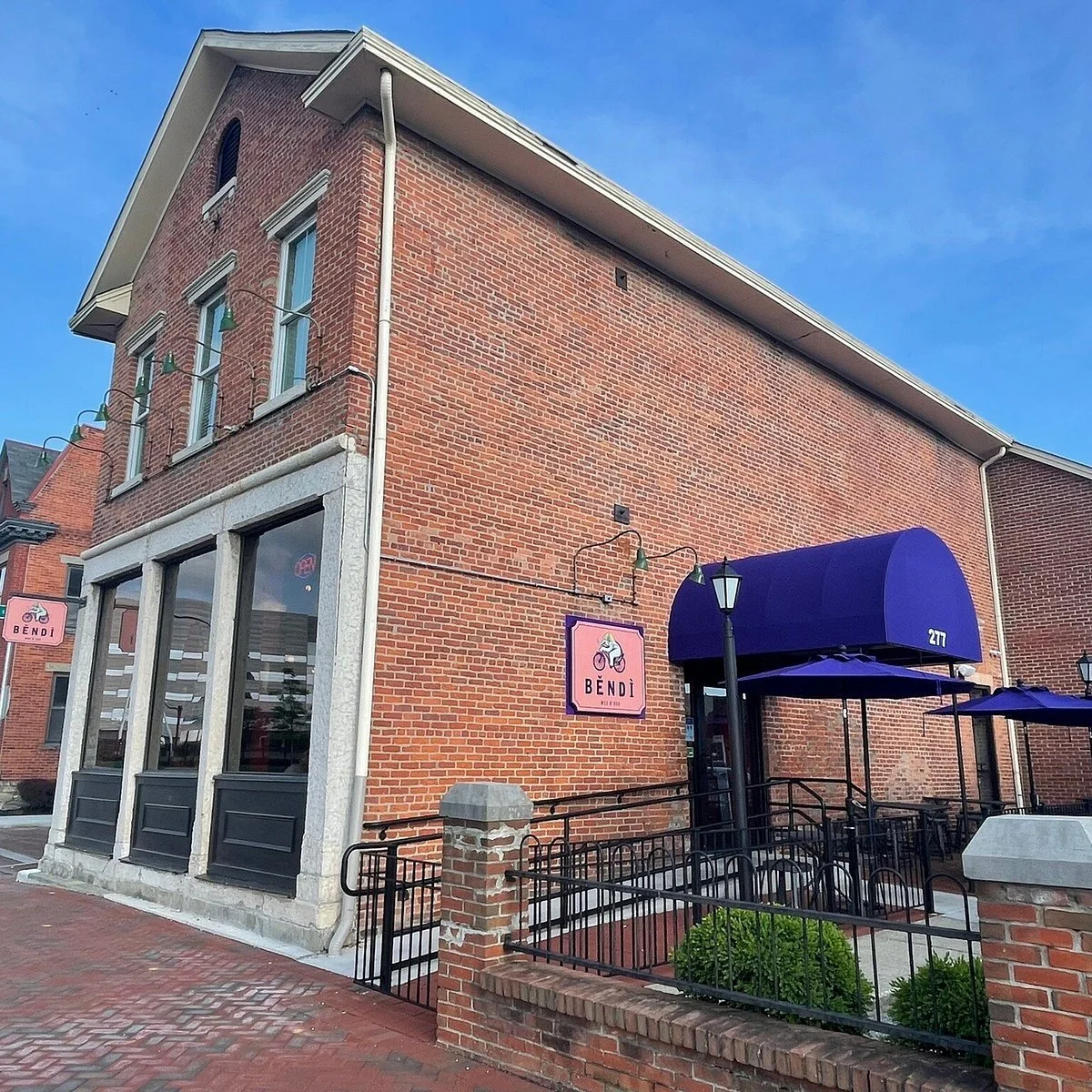 A brick building with a restaurant named BENDI, featuring large windows, a purple awning, outdoor seating area with purple umbrellas, decorative street lamps, and a sidewalk.