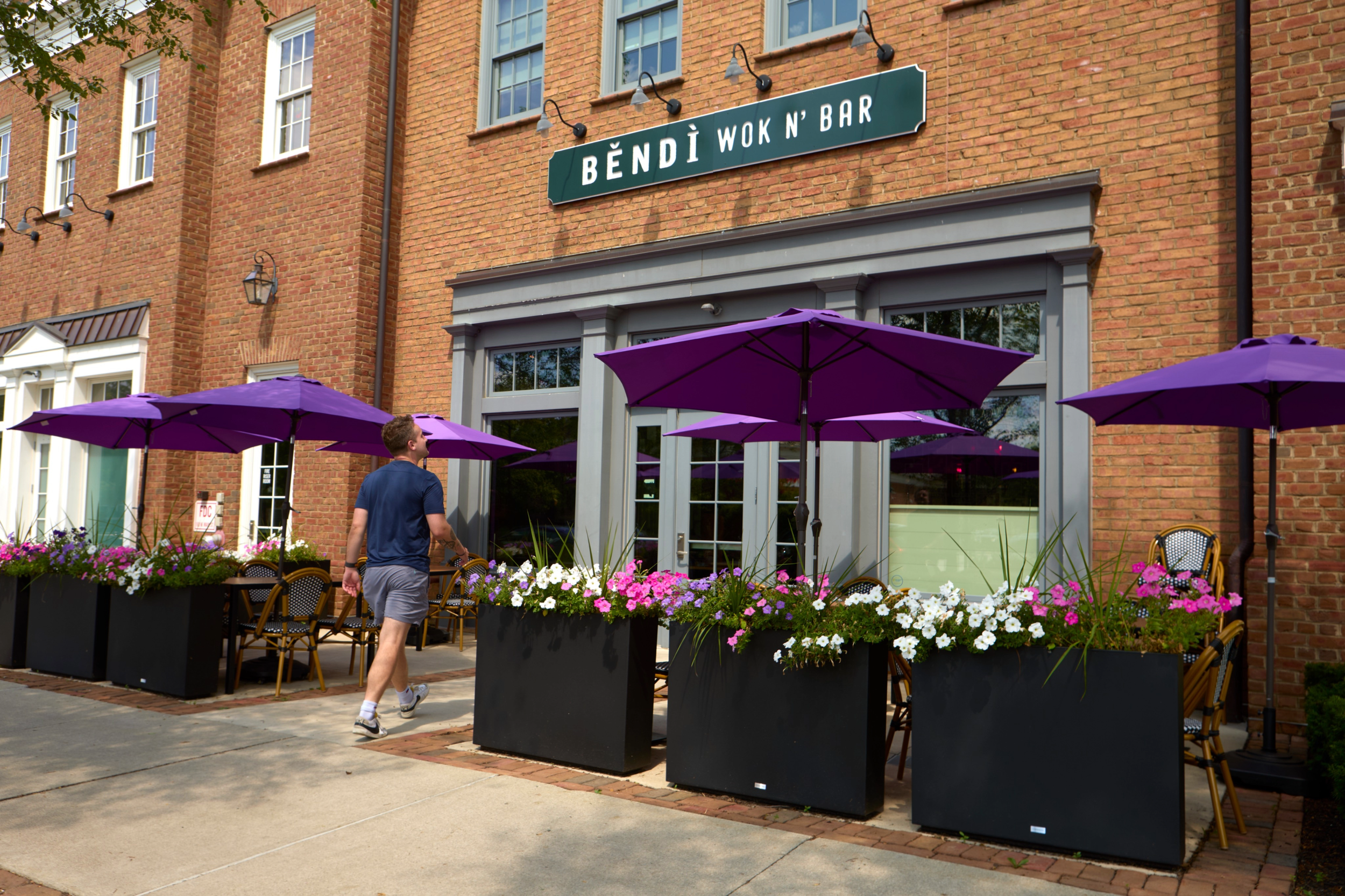 The photo depicts the exterior of a brick building with signage that reads 'BENDI WOK N' BAR.' There are several large purple umbrellas set up outside, and colorful flower planters with pink, white, and purple flowers are arranged along the sidewalk. A man in a blue shirt and shorts walks along the sidewalk, with outdoor seating visible.