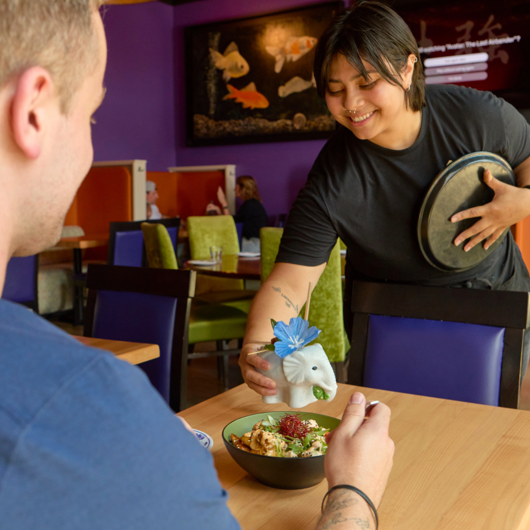 A server places a decorative elephant-shaped condiment holder with a blue flower on a bowl of food for a customer in a restaurant with colorful chairs and aquatic-themed wall art.