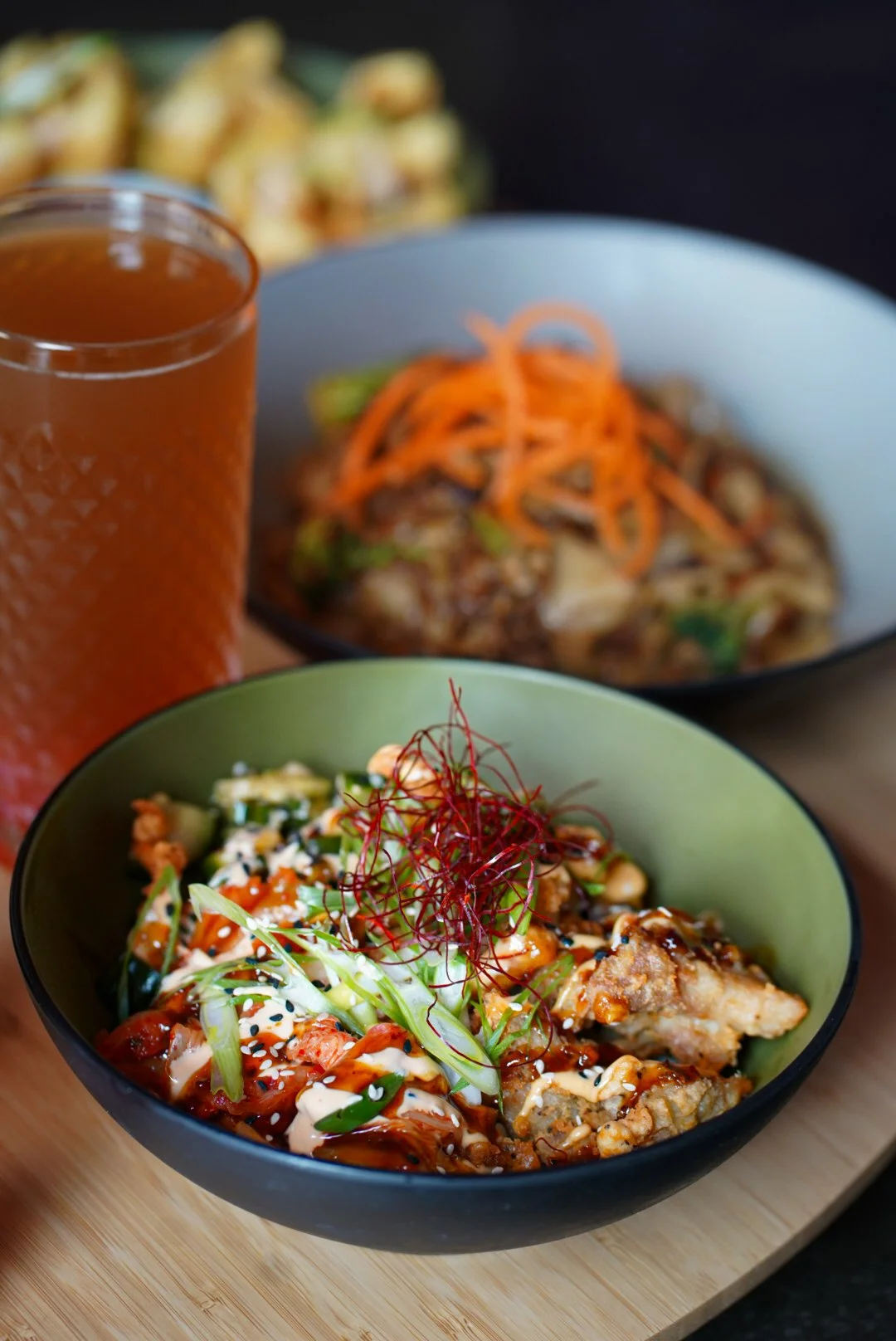 A bowl of Asian-style salad with vegetables, sesame seeds, and crispy chicken, garnished with thin red chili threads and green onion, placed on a wooden board. In the background, there are bowls with noodles topped with shredded carrots, and a glass of iced tea or juice.