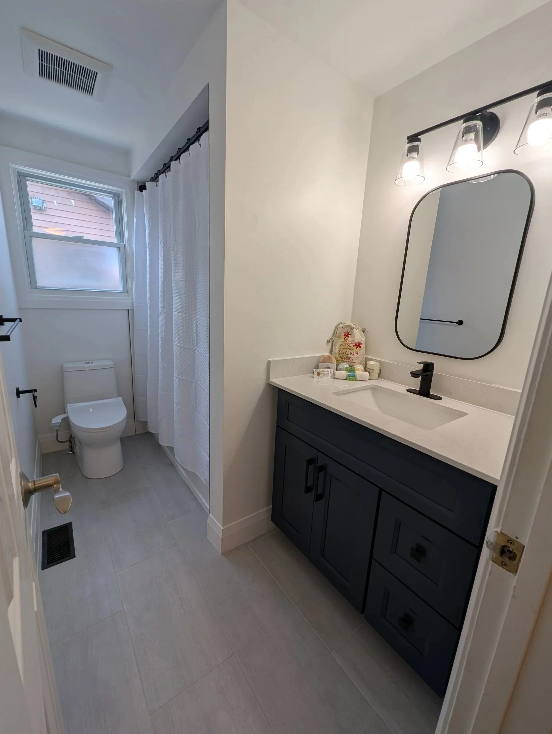 A small bathroom with light gray tiled floor, white walls, and a window with frosted glass. There is a toilet near the window, a white shower curtain on a black rod, and a bathroom vanity with a white countertop, dark gray cabinet, and a black faucet. Above the sink is a rectangular mirror with rounded edges and a black frame, and three light bulbs in glass fixtures are mounted on a black bar above the mirror.