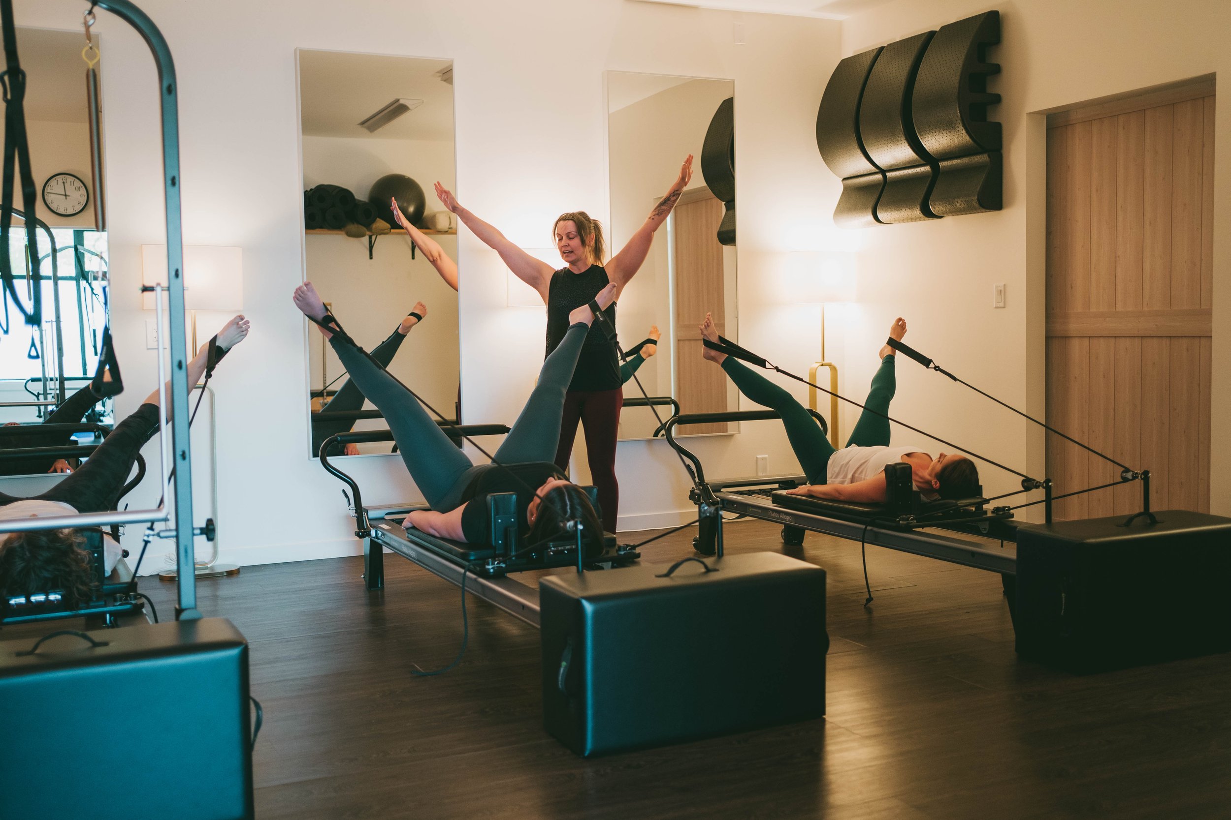 People performing Pilates exercises on reformer machines in a brightly lit fitness studio with mirrors on the wall.
