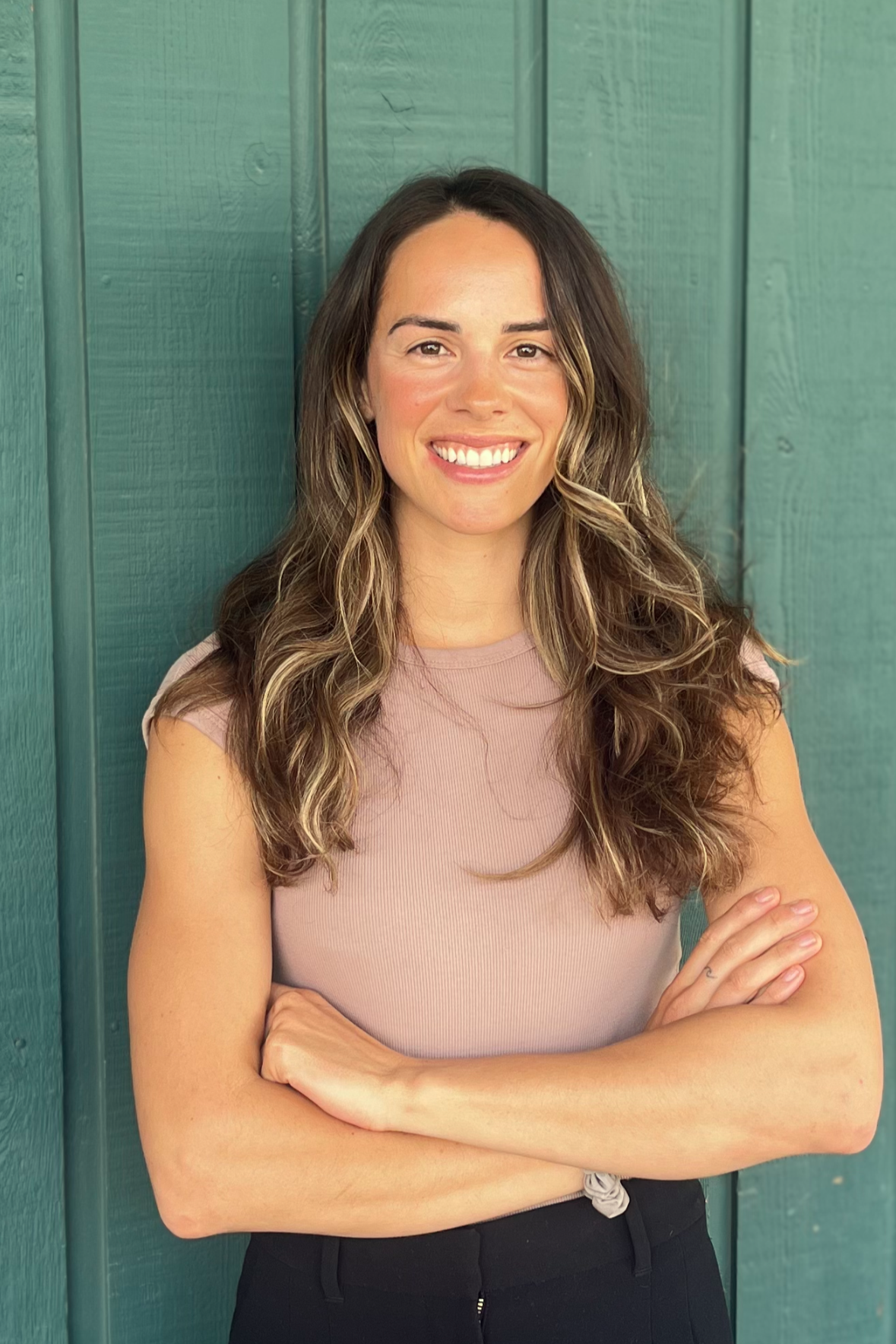 A smiling woman with long brown hair leaning against a green wooden wall, wearing a brown cardigan.