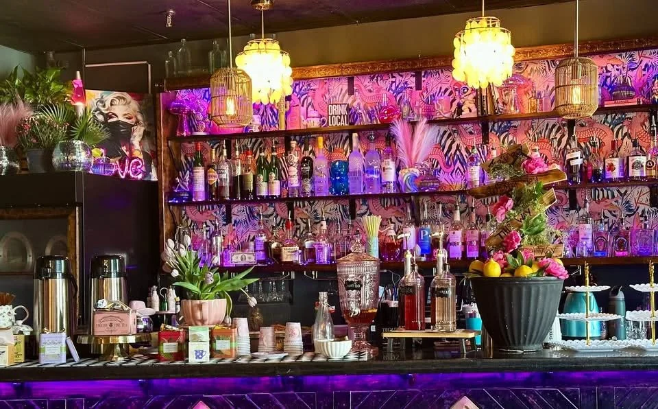 Colorful bar setup with bottles, plants, and decorations on shelves, illuminated by warm and neon lights. A floral arrangement and coffee dispensers are on the counter. Baby shower, wedding shower, wedding, celebration, private dining, private events