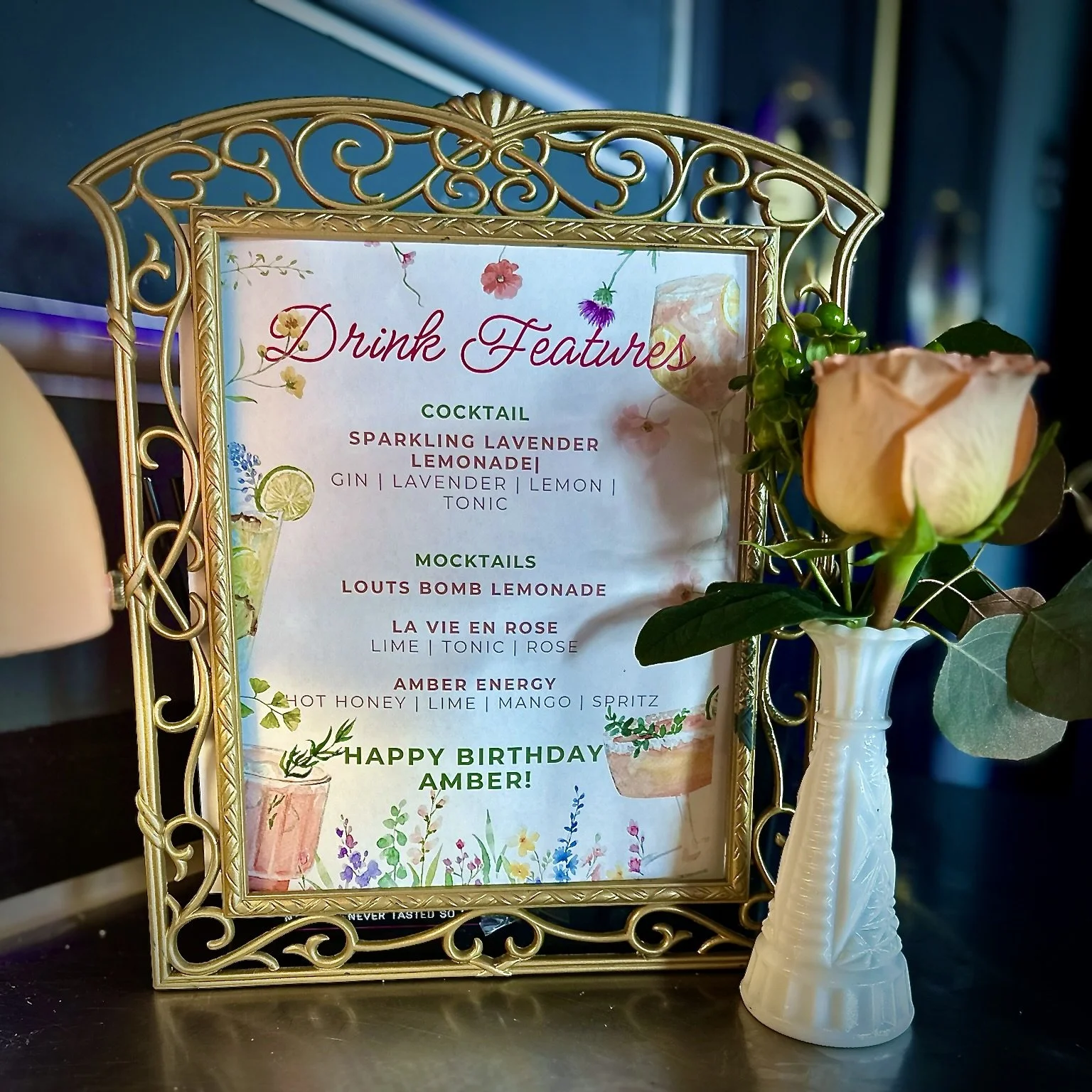 A framed menu titled "Drink Features" listing cocktails and mocktails, including "Sparkling Lavender Lemonade," "Louts Bomb Lemonade," "La Vie en Rose," and "Amber Energy." A rose in a vase is placed next to the frame, and the message "Happy Birthday