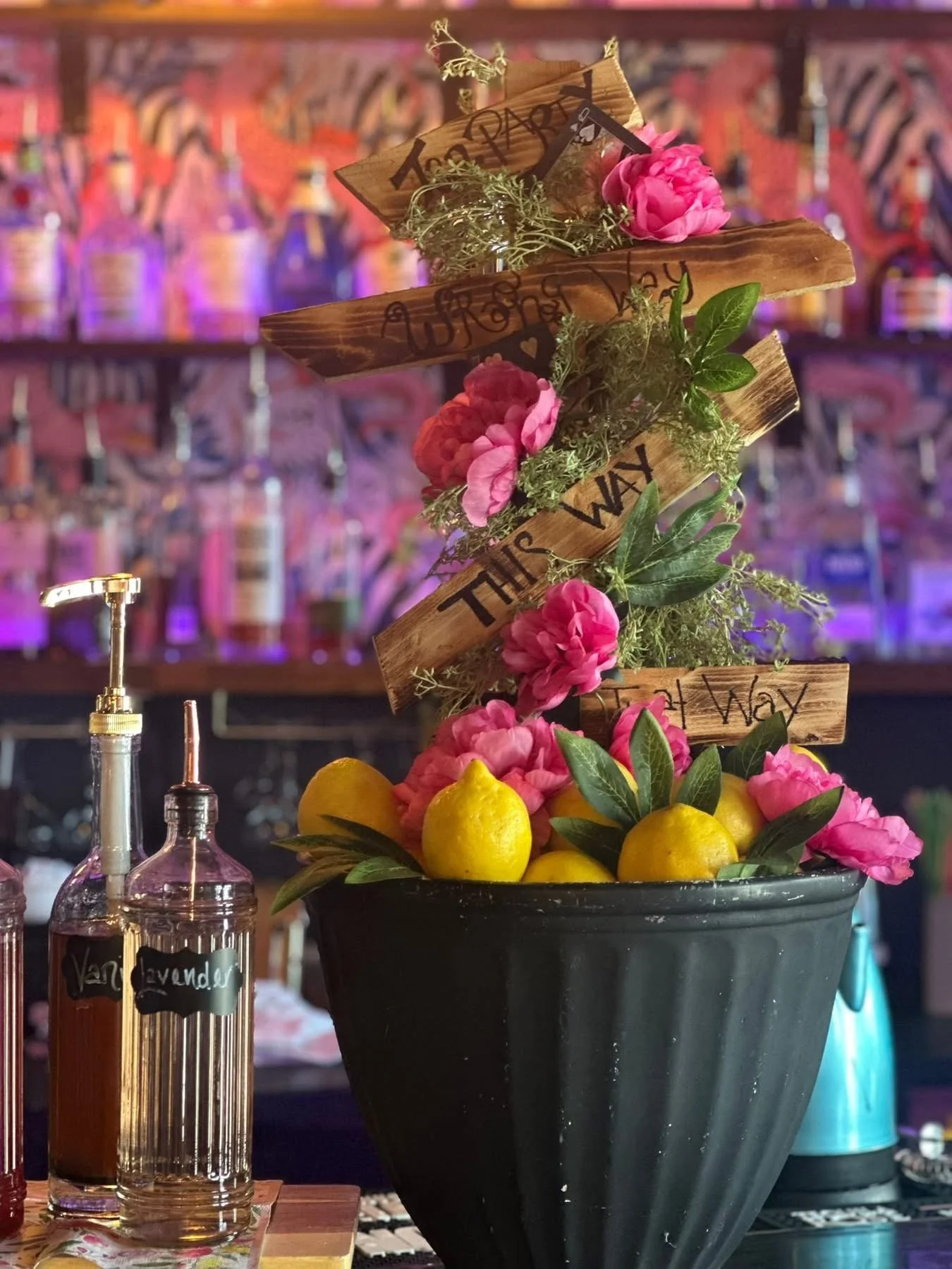 Decorative setup with a black pot filled with lemons and pink flowers, surrounded by wooden signs with writing, on a bar counter with bottles in the background. Tea Party Pop up Event,  mothers day, Baby shower, wedding shower, wedding, celebration, 