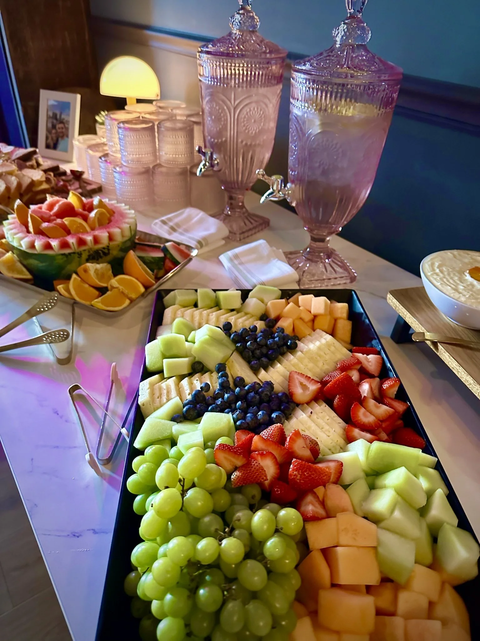 Assorted fresh cut fruits on a black rectangular platter, including green grapes, pineapple, strawberries, cantaloupe, honeydew, blueberries, and watermelon. Shower, Celebration, Event, Private Rental, Party, Micro Wedding. 