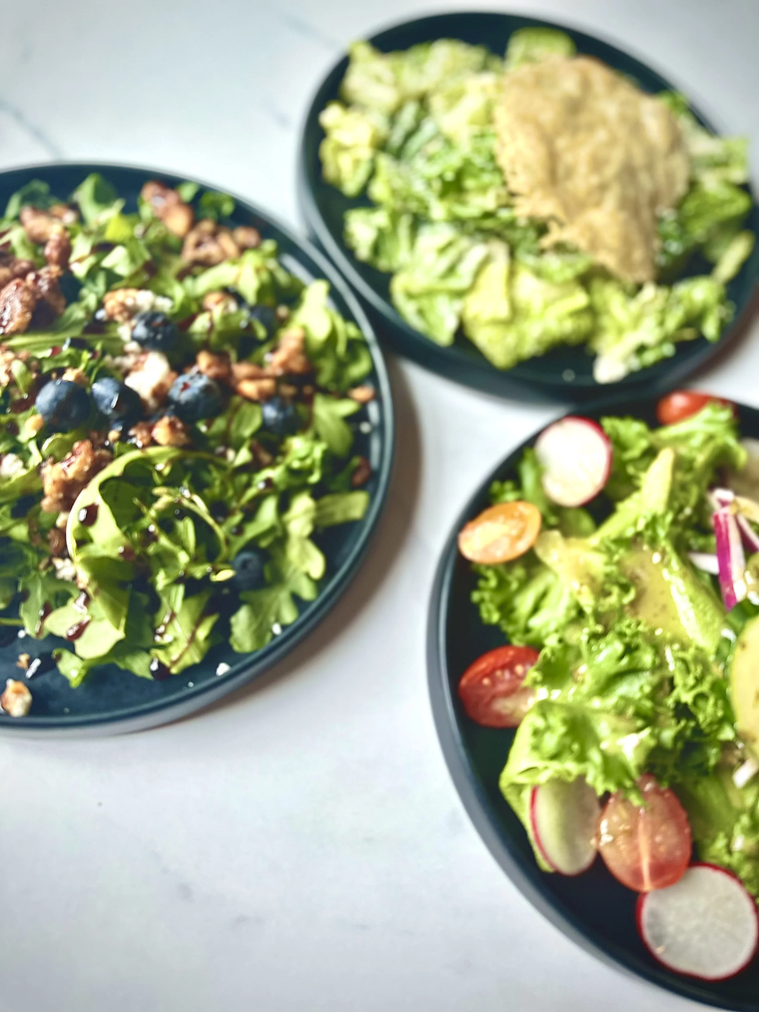 Three plates of salads on a white table. The first salad features mixed greens with blueberries, walnuts, and cheese. The second plate contains a Caesar salad with lettuce and a parmesan crisp on top. The third salad includes lettuce, sliced radishes