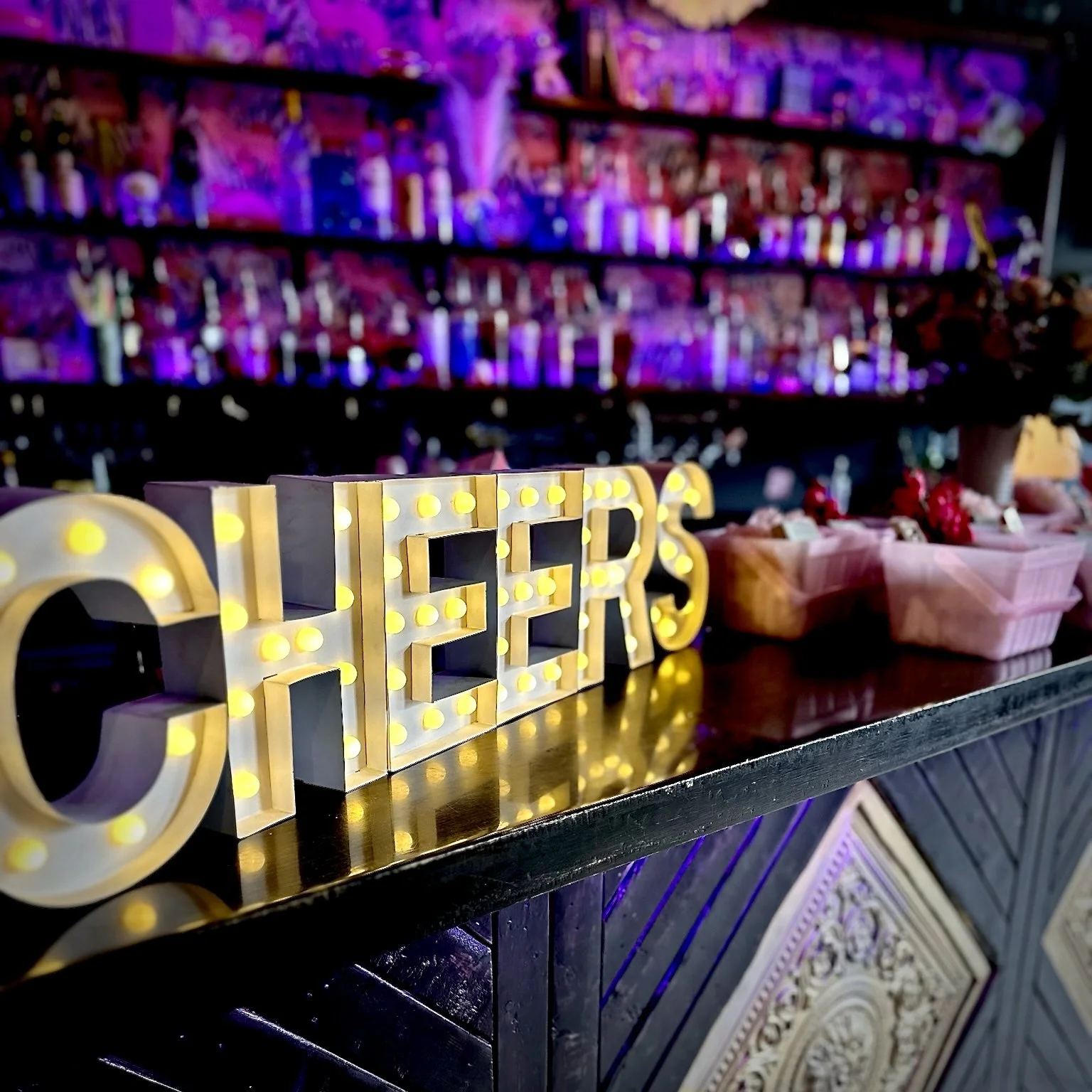 Illuminated sign spelling 'CHEERS' on a bar countertop with beverages and colorful bottles in the background. Shower, Celebration, Event, Private Rental, Party, Micro Wedding.  Birthday Party.