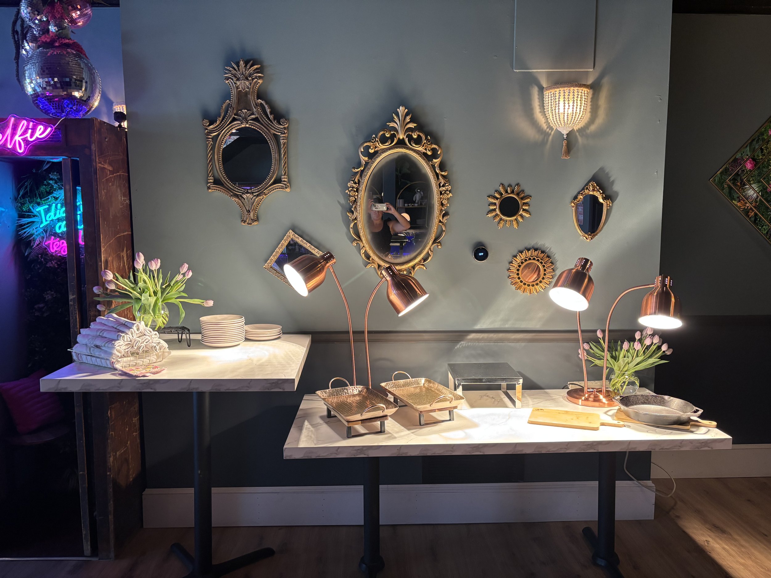 A decorative display with a collection of ornate mirrors on a wall, two tables with white surfaces, copper lamps, stacked plates, folded napkins, and tulip flower arrangements. The tables hold cookware and cutting boards. A neon sign is visible in th